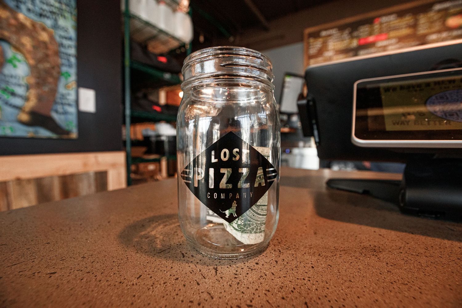 Glass mason jar with 'Lost Pizzas' logo on a wooden counter in a restaurant setting.