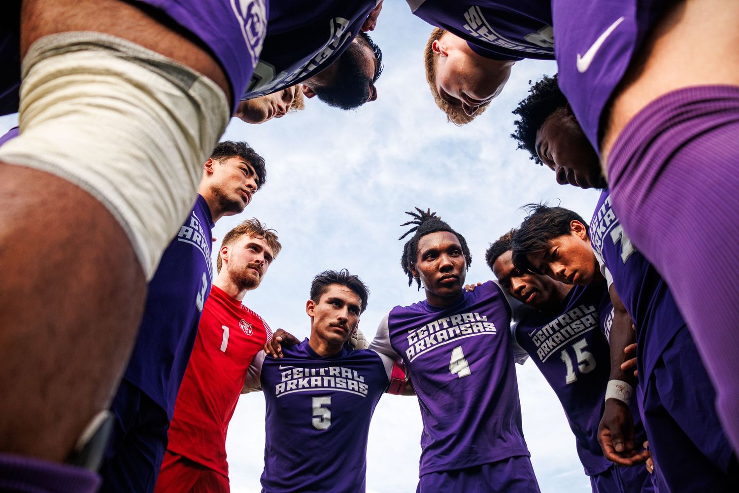 College football teammates huddle together in a circle wearing purple jerseys against blue sky.