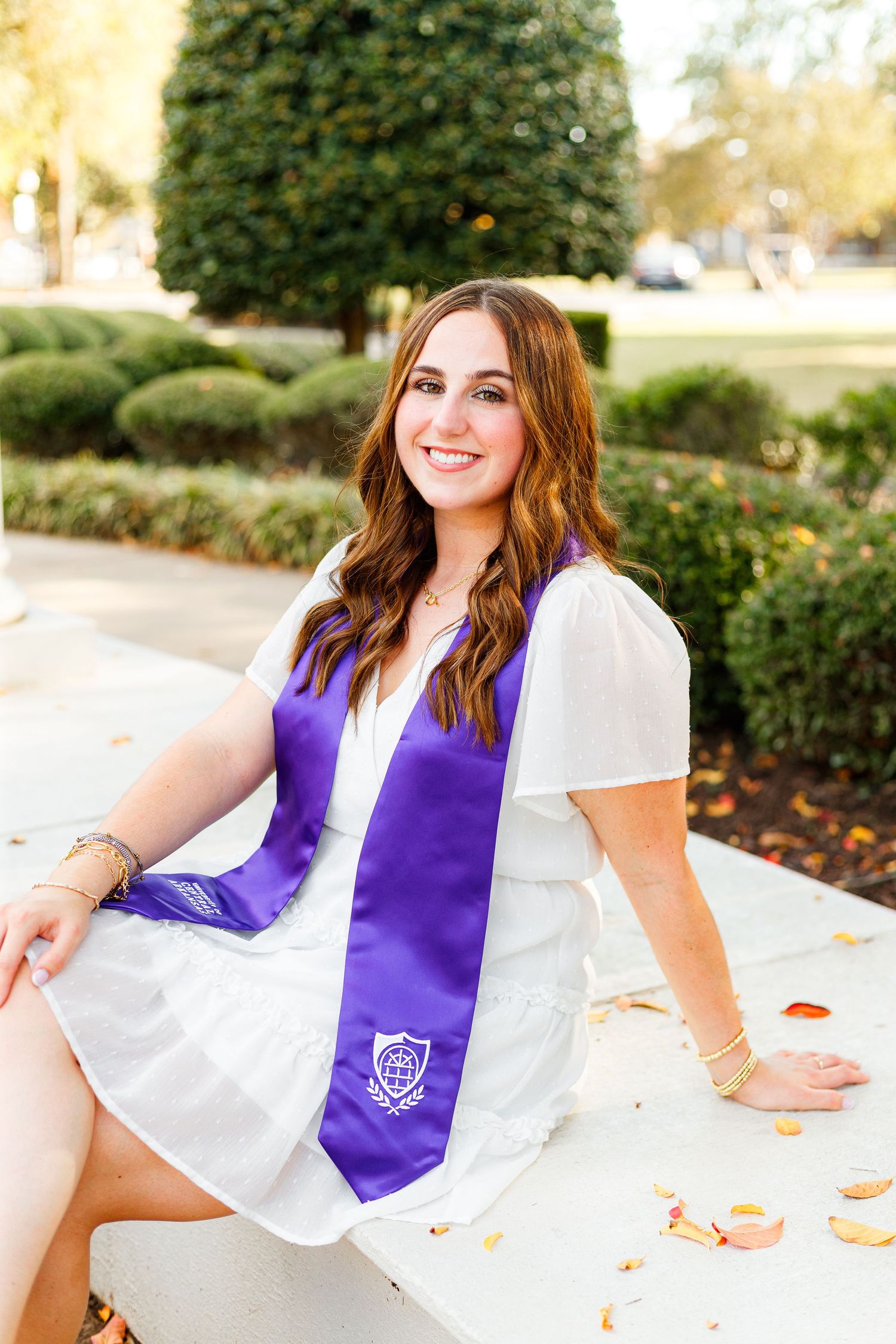 Graduate in white top and purple graduation sash poses outdoors during senior photo session.