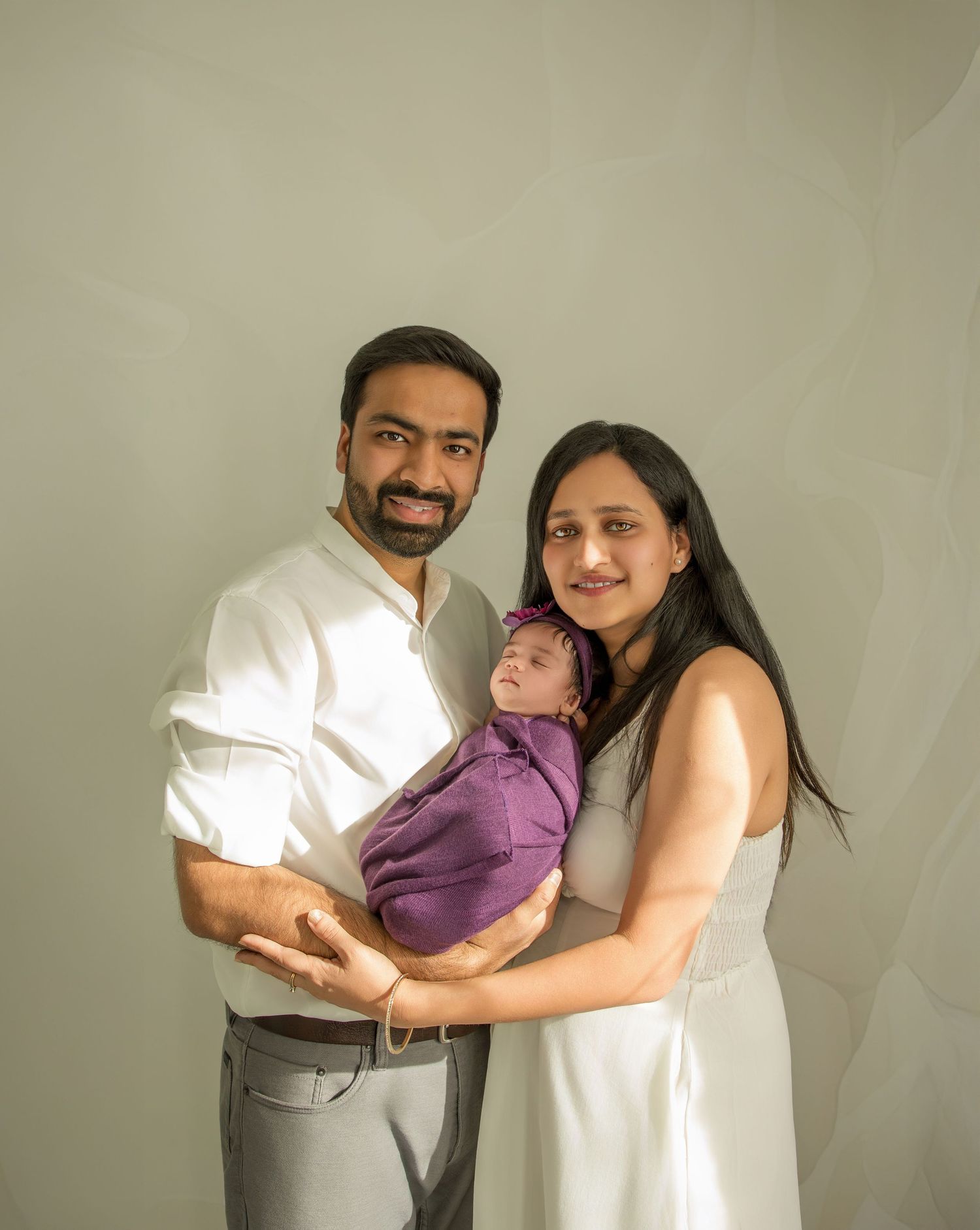 A family of three in white attire poses together against a neutral background while holding their newborn baby in purple.