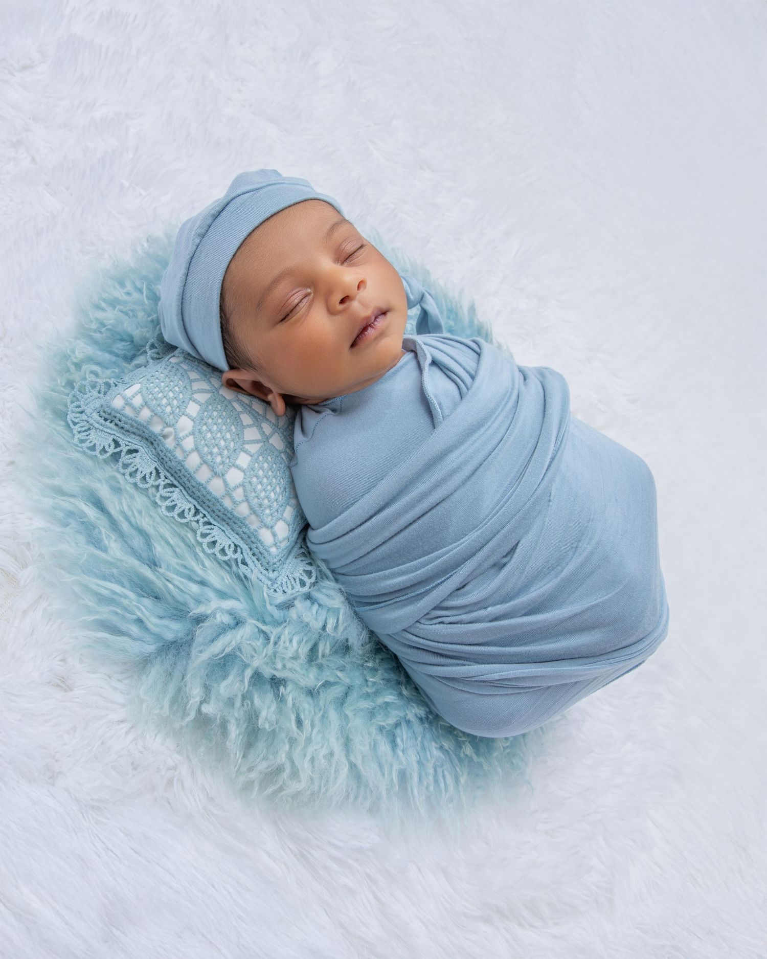 A sleeping newborn baby wrapped in soft blue fabric lies on a lacy white blanket in a professional photography setup.
