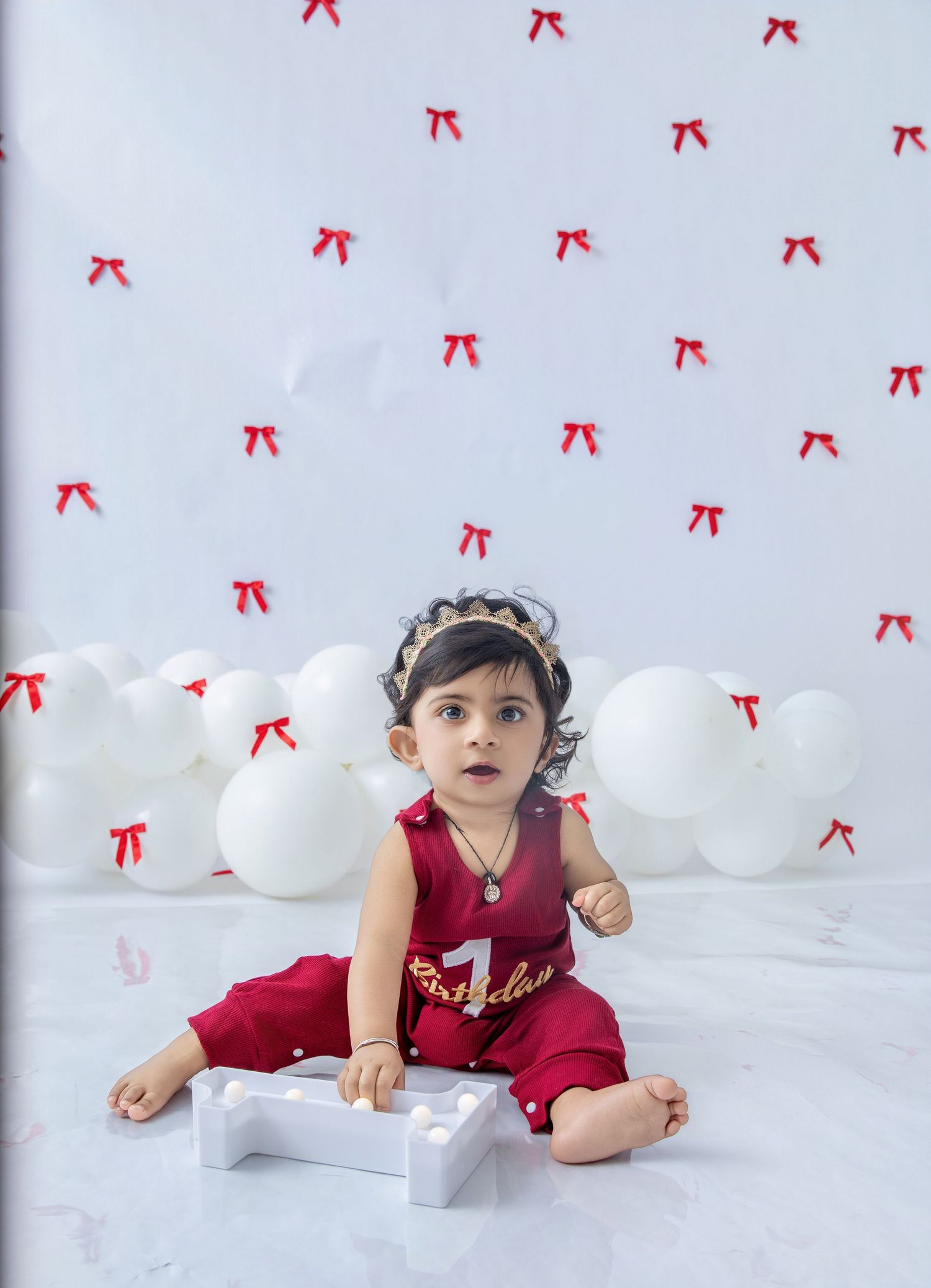 Child in red outfit sits on white floor with decorative number one and balloons.