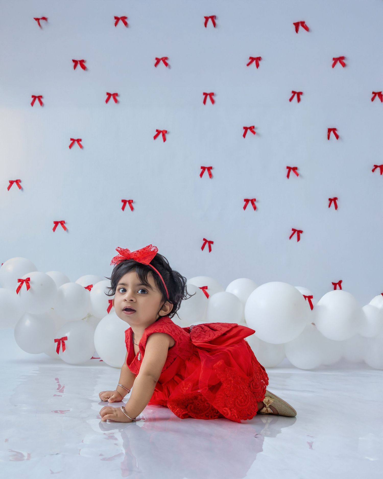 Child in red dress crawling on white floor with floating balloons and red bows.