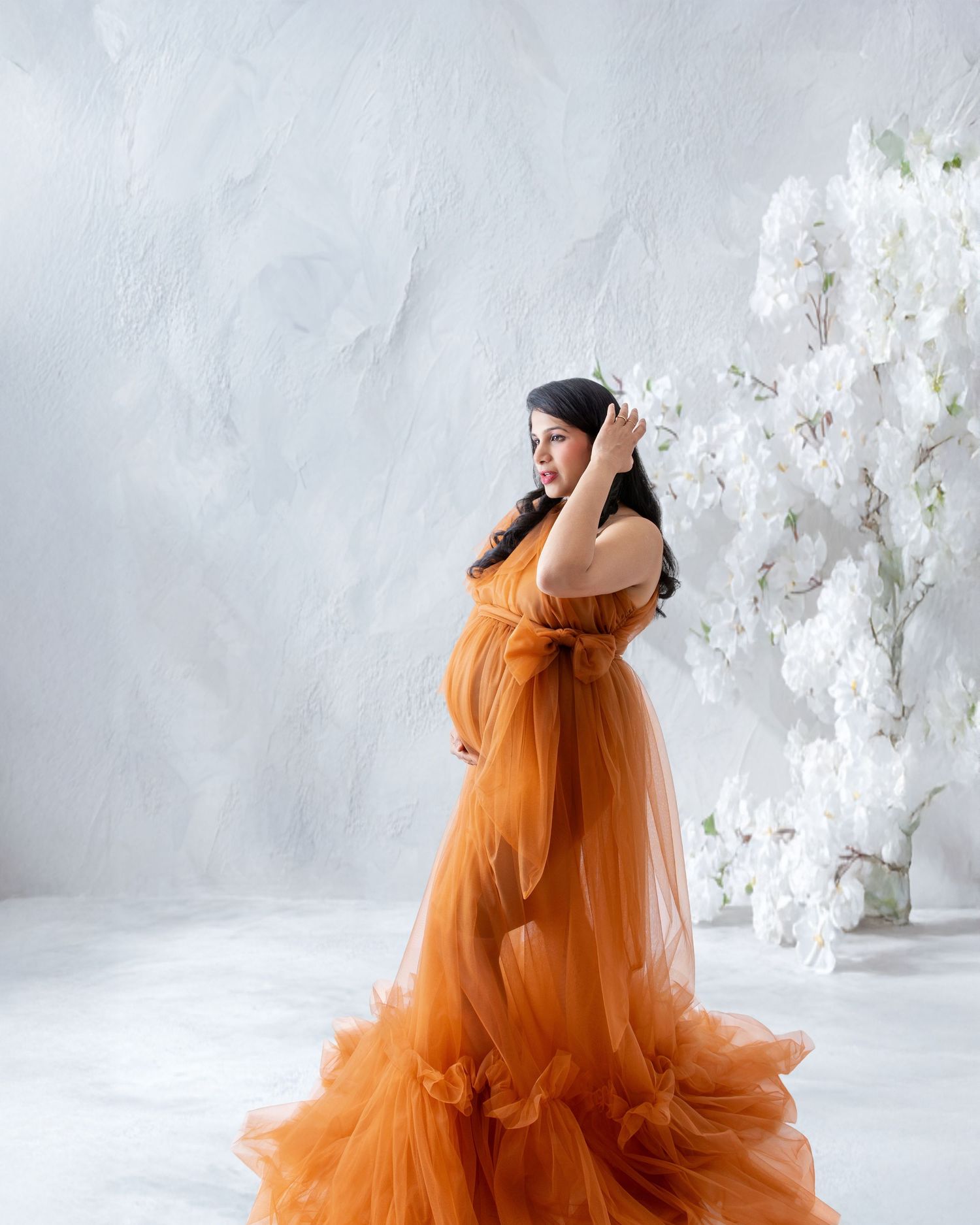 Pregnant woman in elegant orange tulle maternity gown poses gracefully in studio with white floral backdrop.