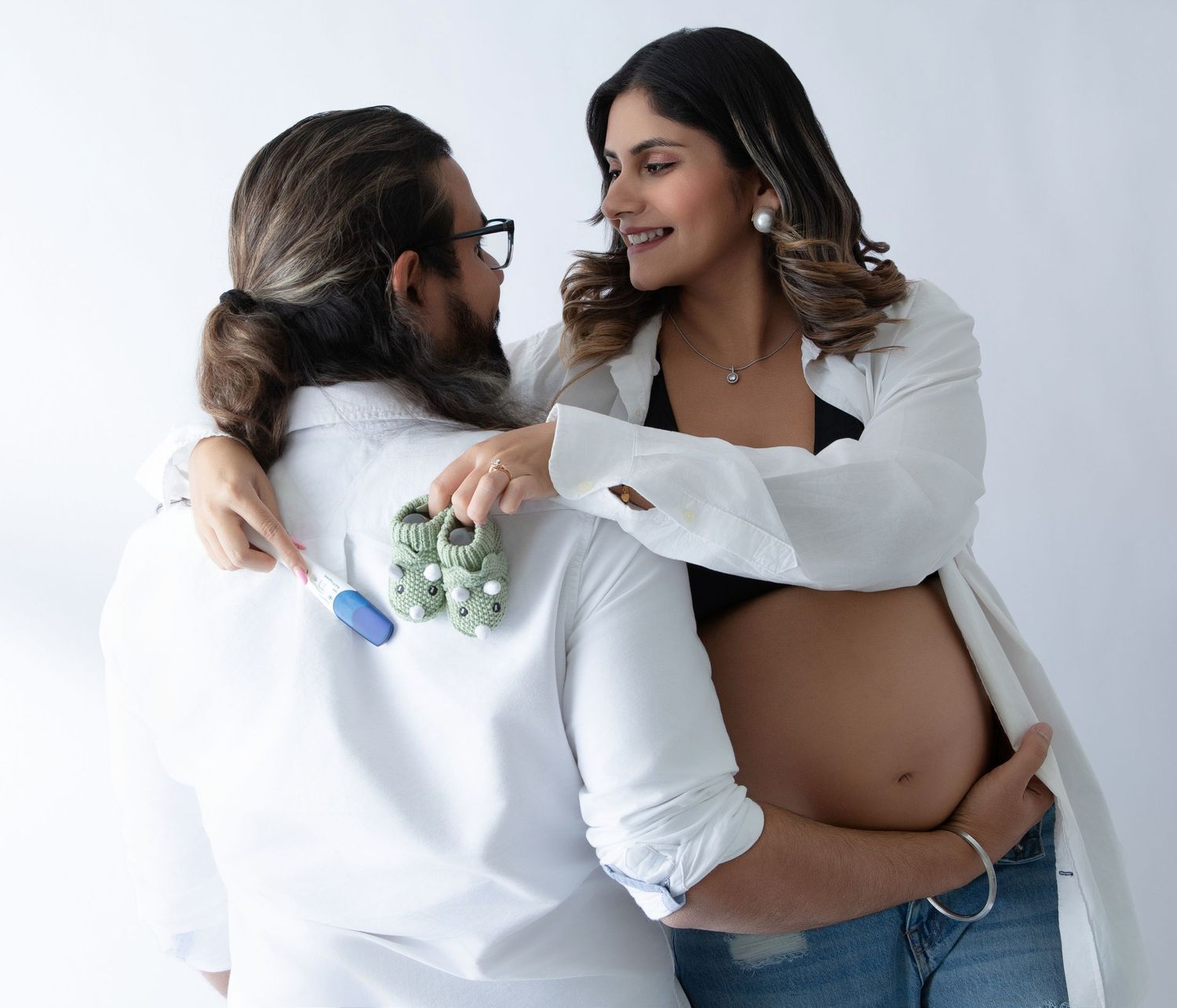 Pregnant woman in white blazer and jeans smiles at partner holding tiny baby booties during maternity photo shoot.