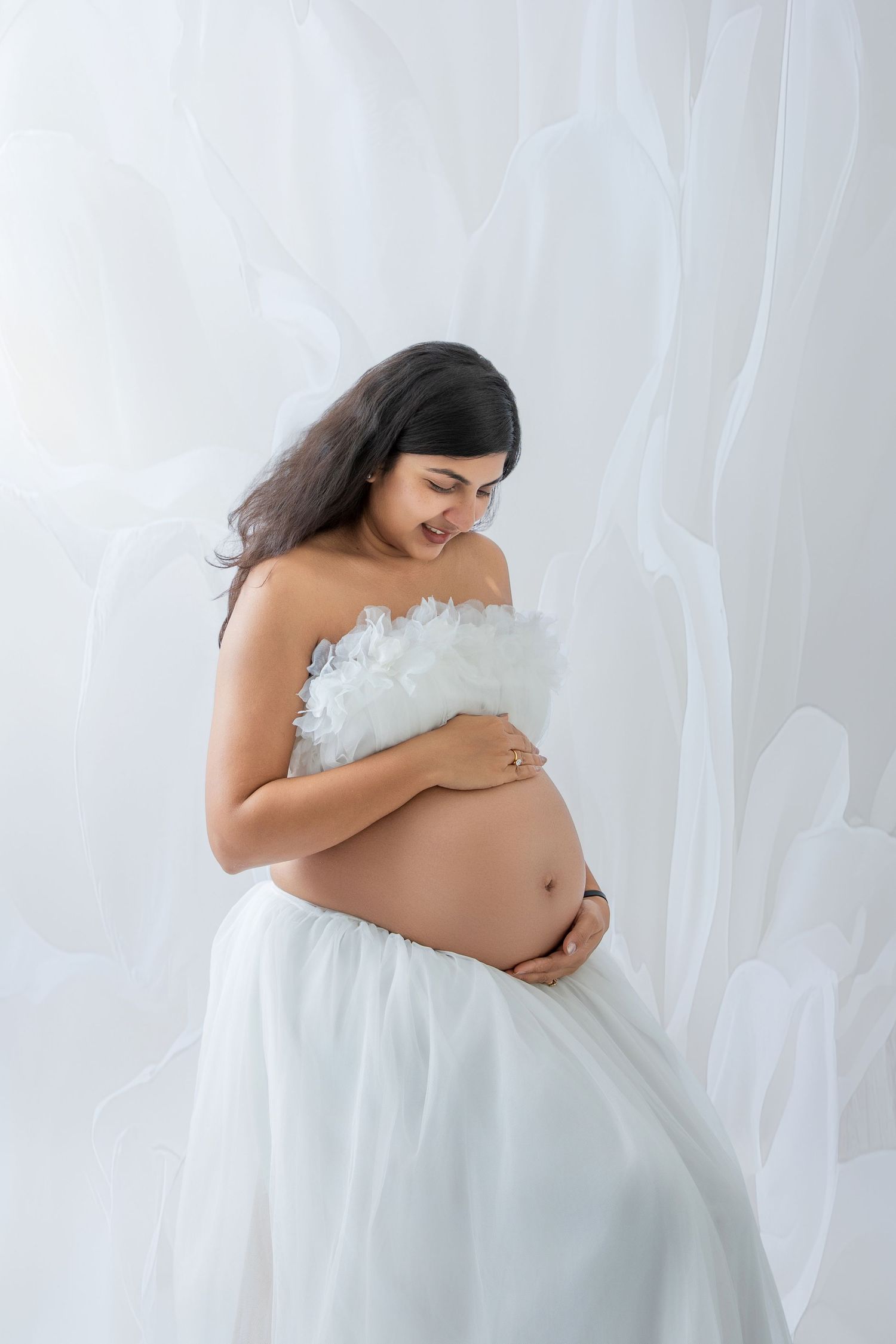 Smiling pregnant woman in white strapless ruffled gown cradling her baby bump against a soft white floral background.