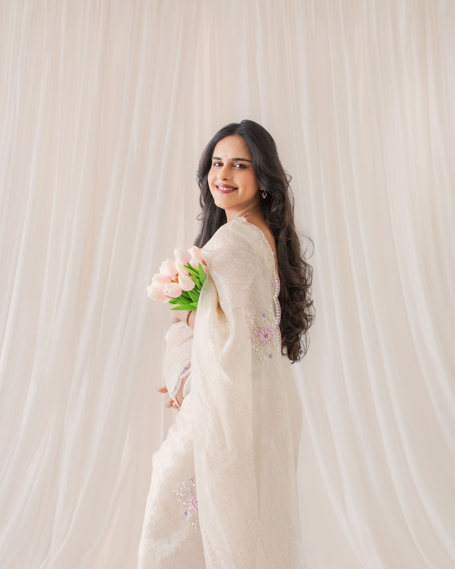 A woman in a white embroidered saree holds pink tulips, smiling gracefully against a soft white curtain backdrop.