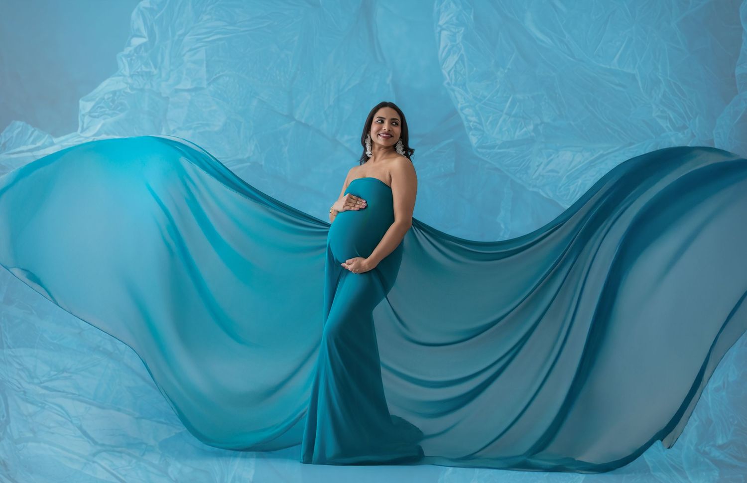 Pregnant woman in flowing teal gown poses gracefully against blue backdrop in elegant maternity photo shoot.