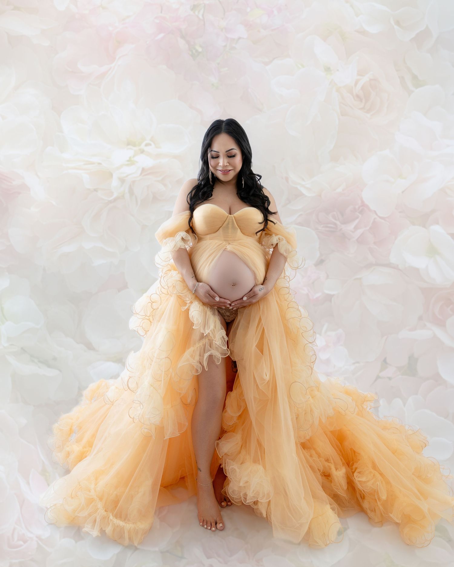 Pregnant woman in elegant peach tulle maternity gown poses gracefully against a soft floral backdrop.