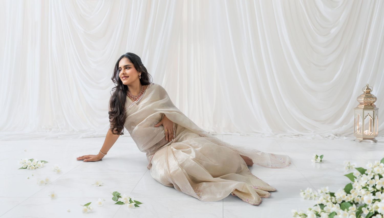 Woman in elegant cream silk saree poses gracefully on white floor with floral decor and draped backdrop.