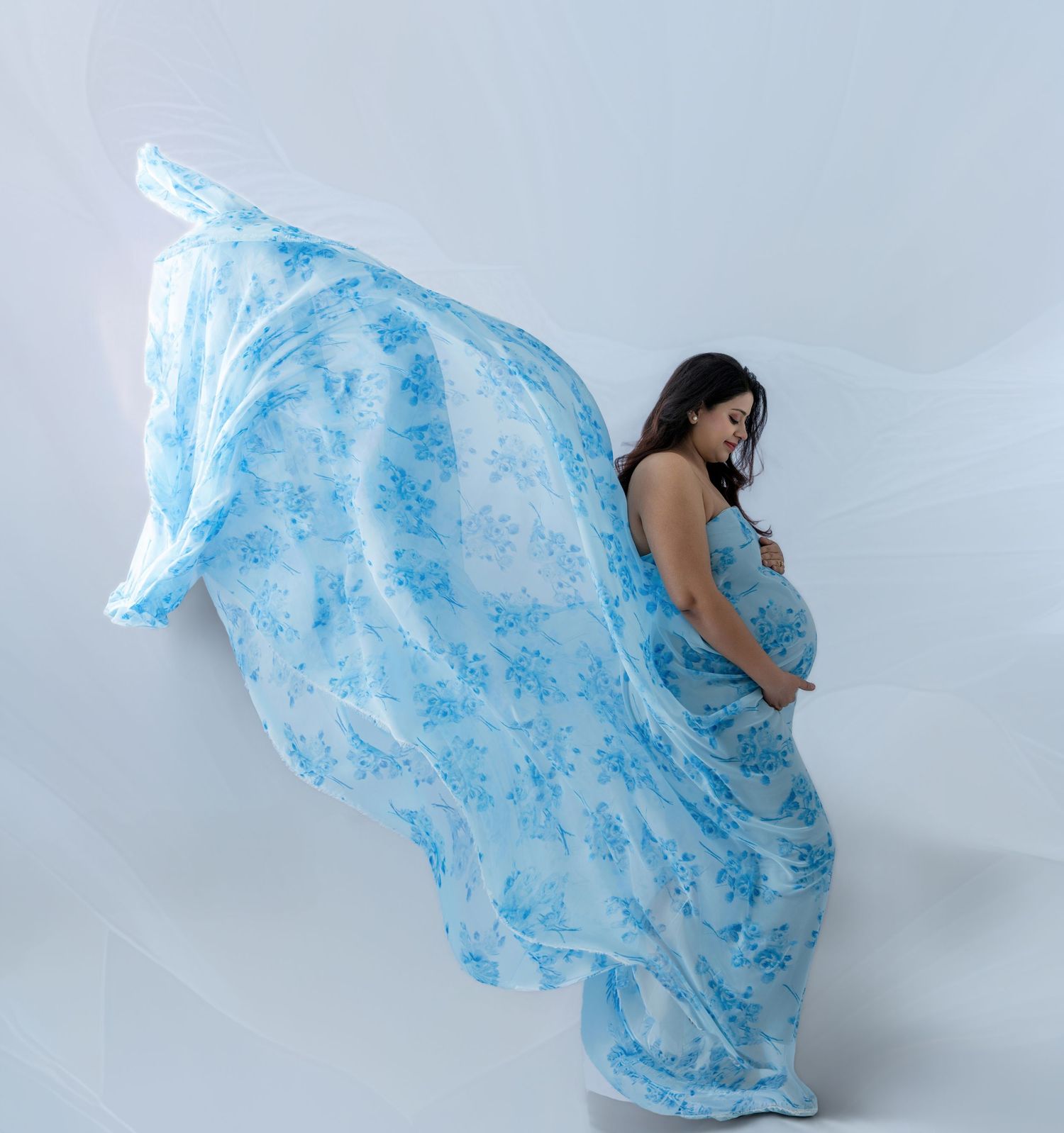 Pregnant woman in flowing blue floral dress cradling baby bump in elegant maternity photo shoot.