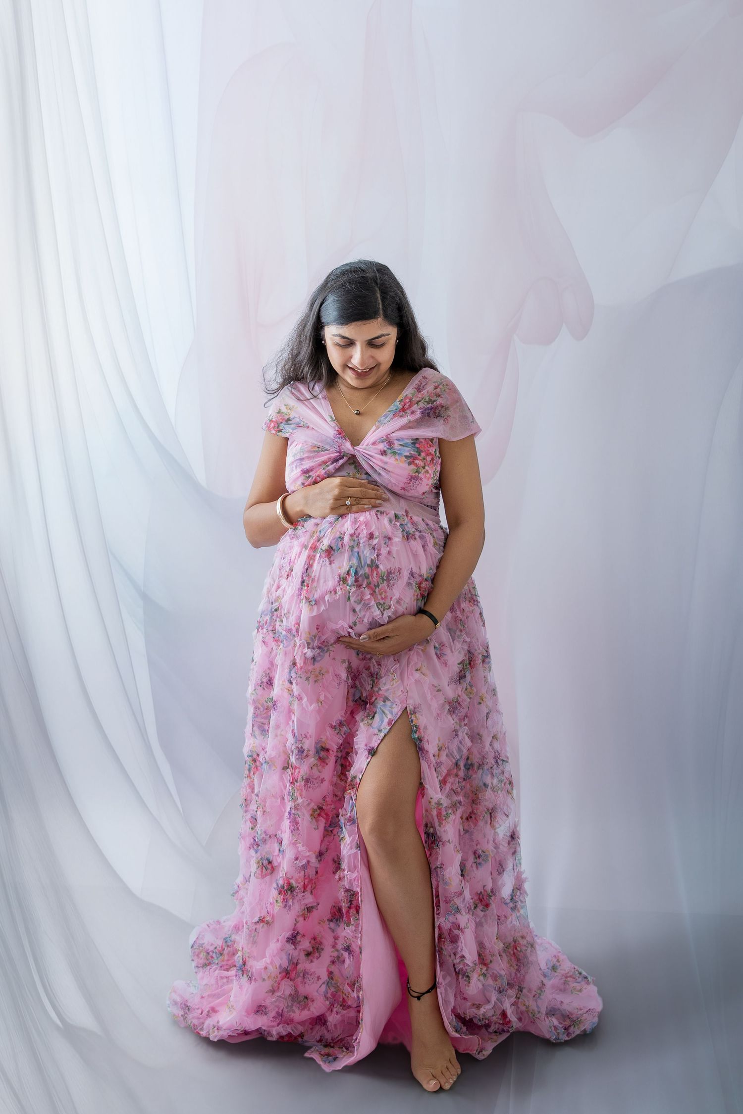 Pregnant woman in pink floral maternity gown cradling baby bump, studio portrait with soft white draped backdrop.