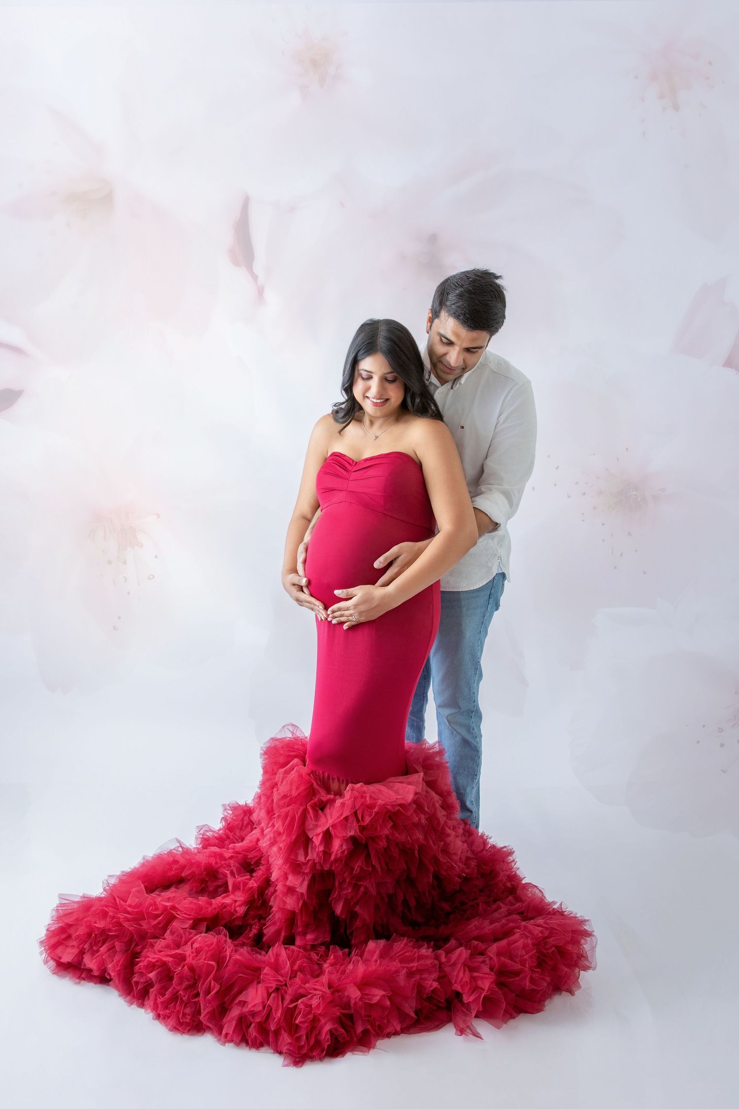 Pregnant woman in red ruffled gown with partner embracing her from behind in a floral studio setting.