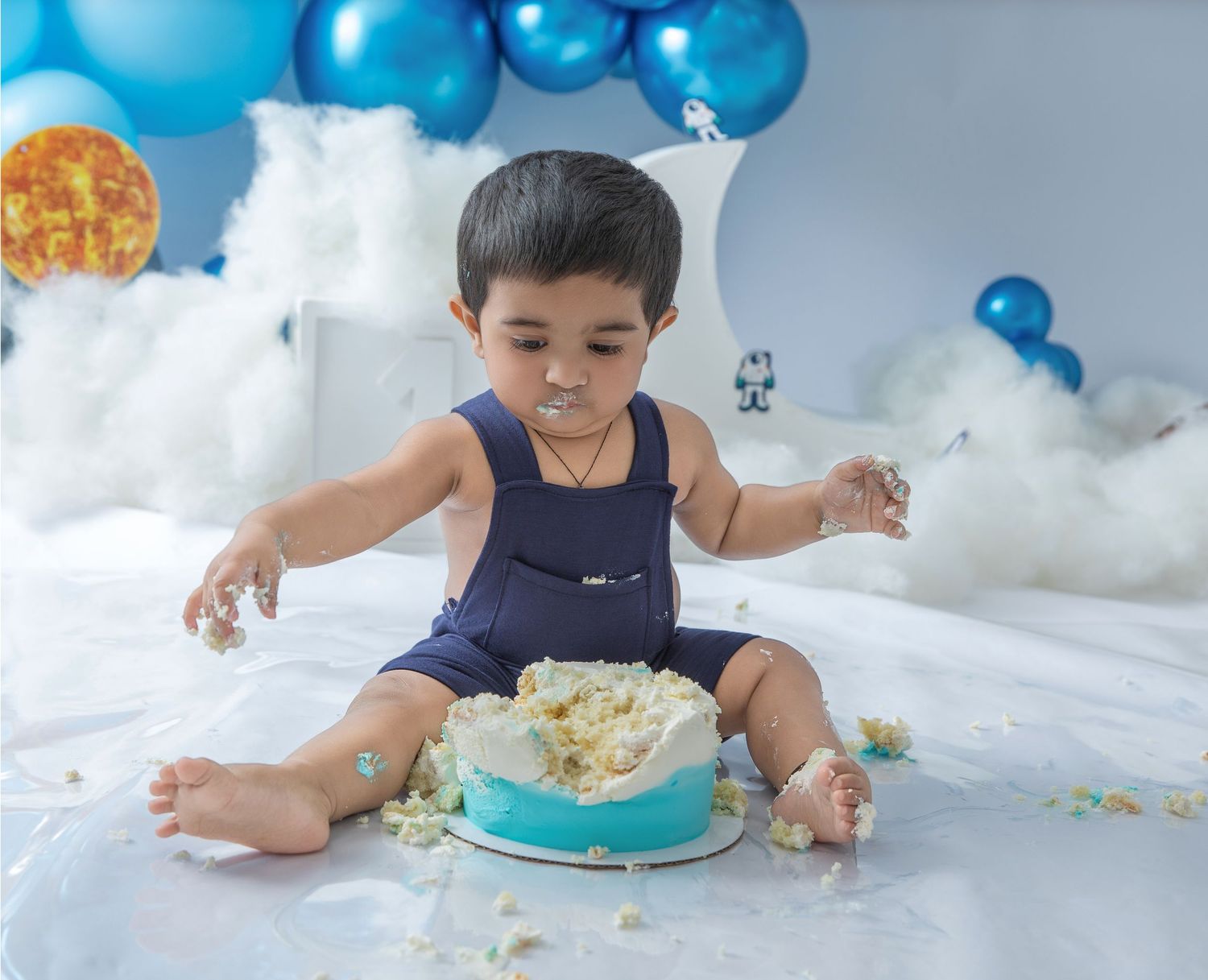 A baby smashing a blue birthday cake during a cake smash photo session with white and blue balloon decorations.