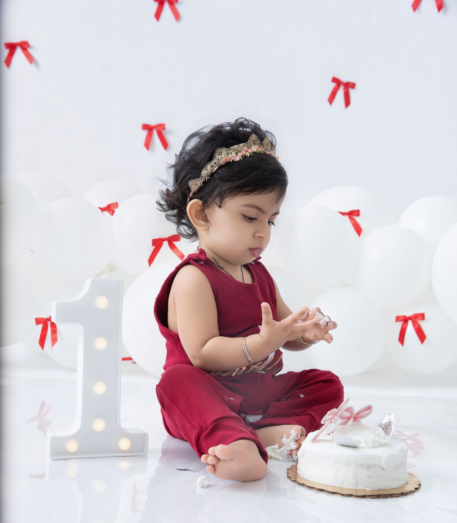 Baby in burgundy dress examining white birthday cake during photo session.