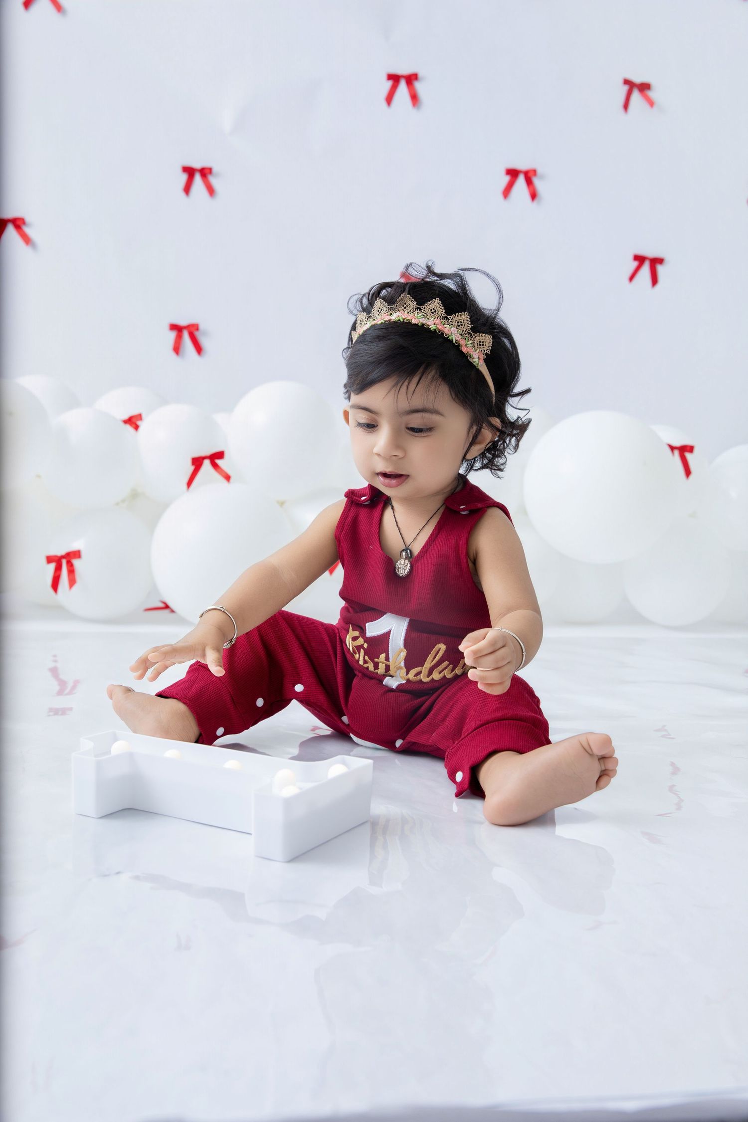 Baby in burgundy outfit exploring white decorated space with crown accessory.