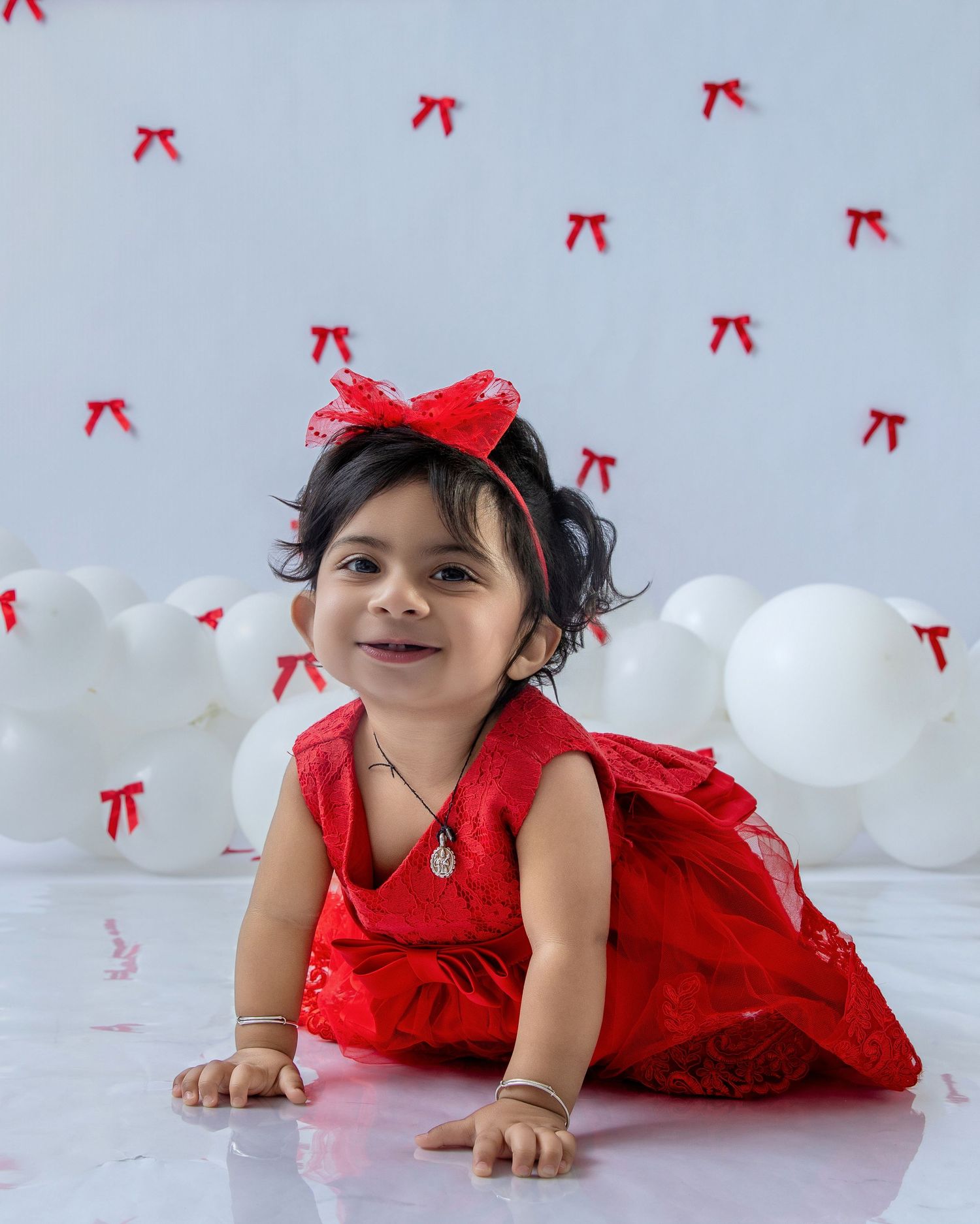 A small child in a festive red dress and bow poses on the floor with white balloons and red bows in the background.