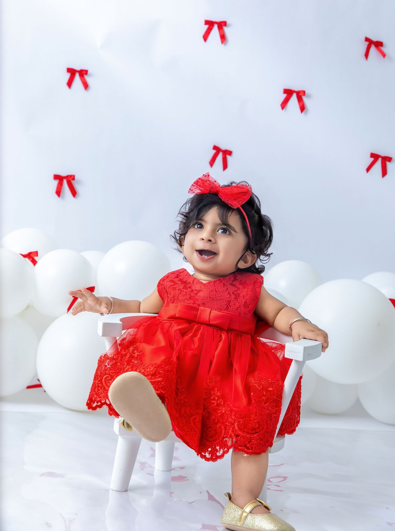 Child in red lace dress playing among white balloons with red bows.