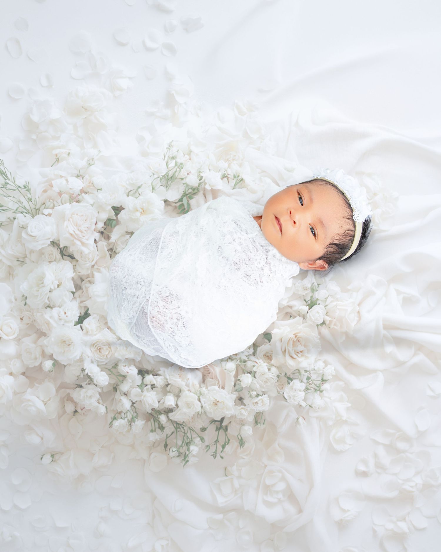 Newborn baby in white lace swaddle and bonnet surrounded by fresh white roses and baby's breath flowers.