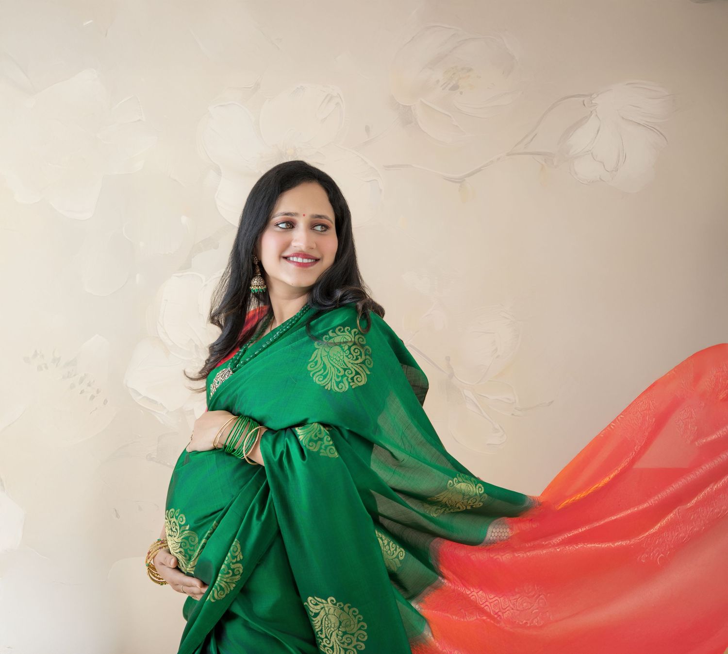 Pregnant woman in elegant green and gold saree with orange drape posing gracefully against a neutral wall.