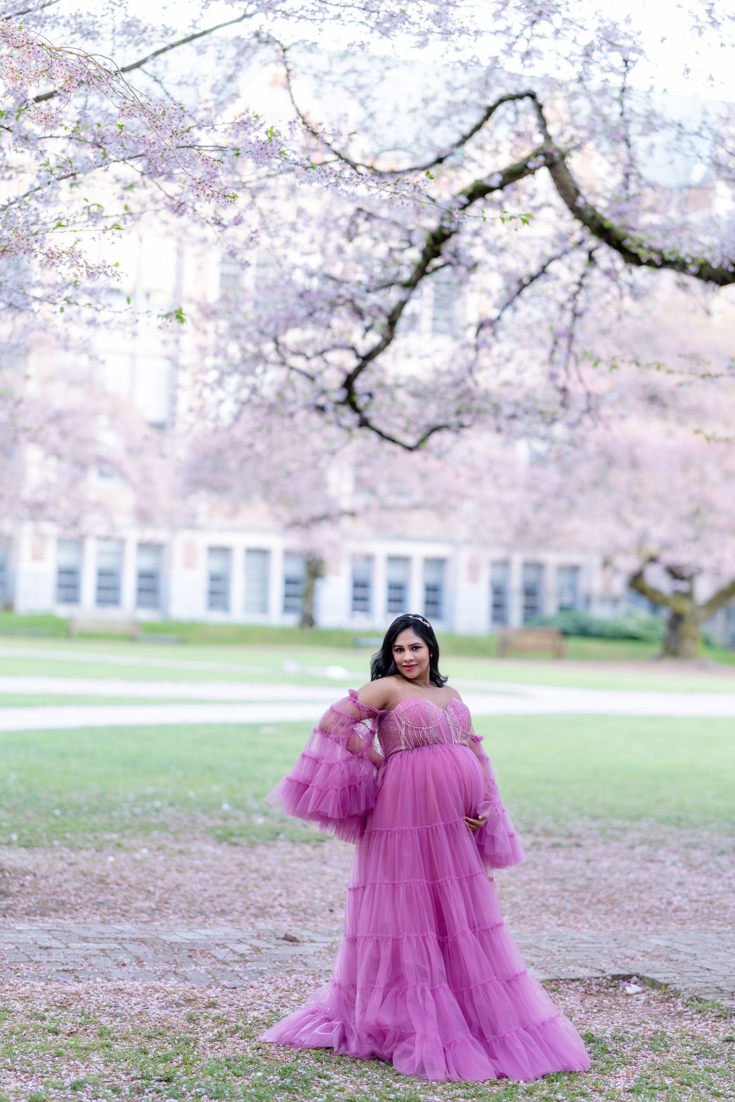 Smiling pregnant woman in a purple tulle off-shoulder gown standing under cherry blossom trees in a park.