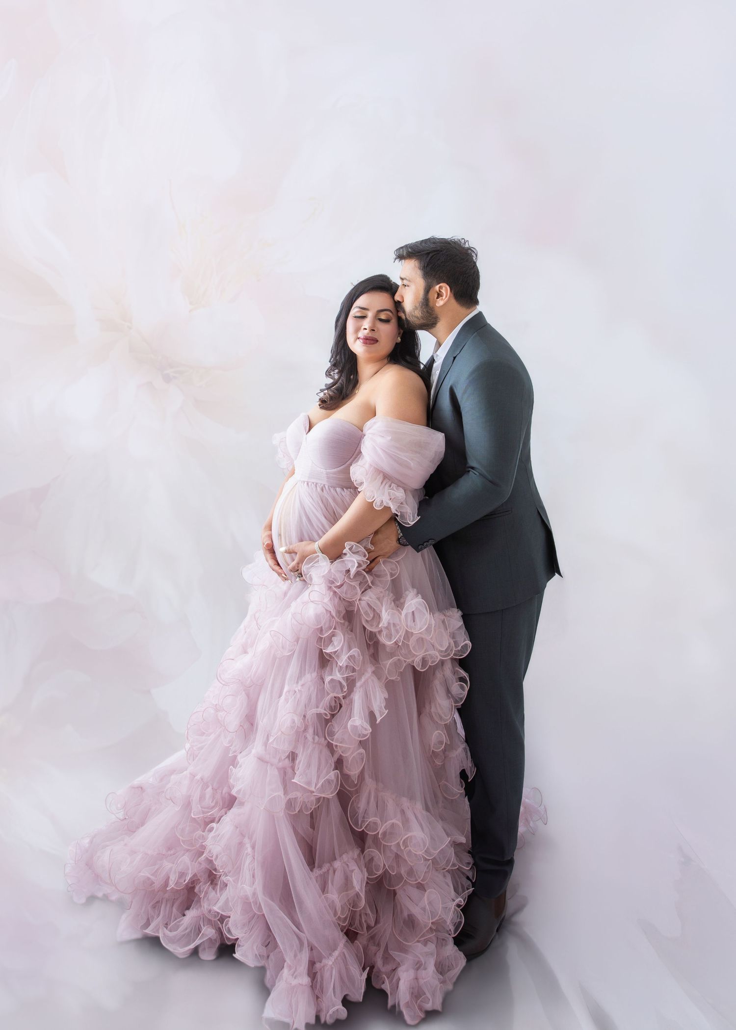 Couple in elegant maternity photo shoot, woman in pink ruffled gown, man in gray suit, soft marble background.