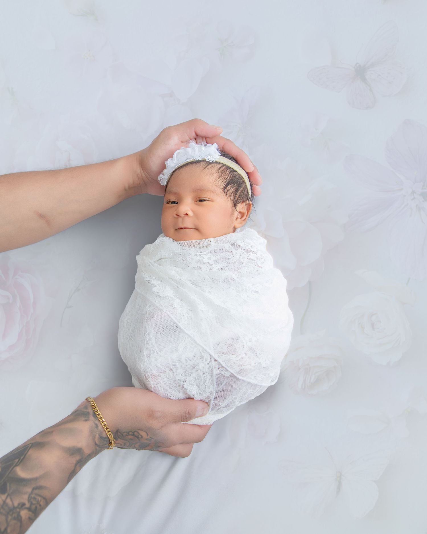 Newborn baby in white lace swaddle and headband gently held by tattooed parent hands.