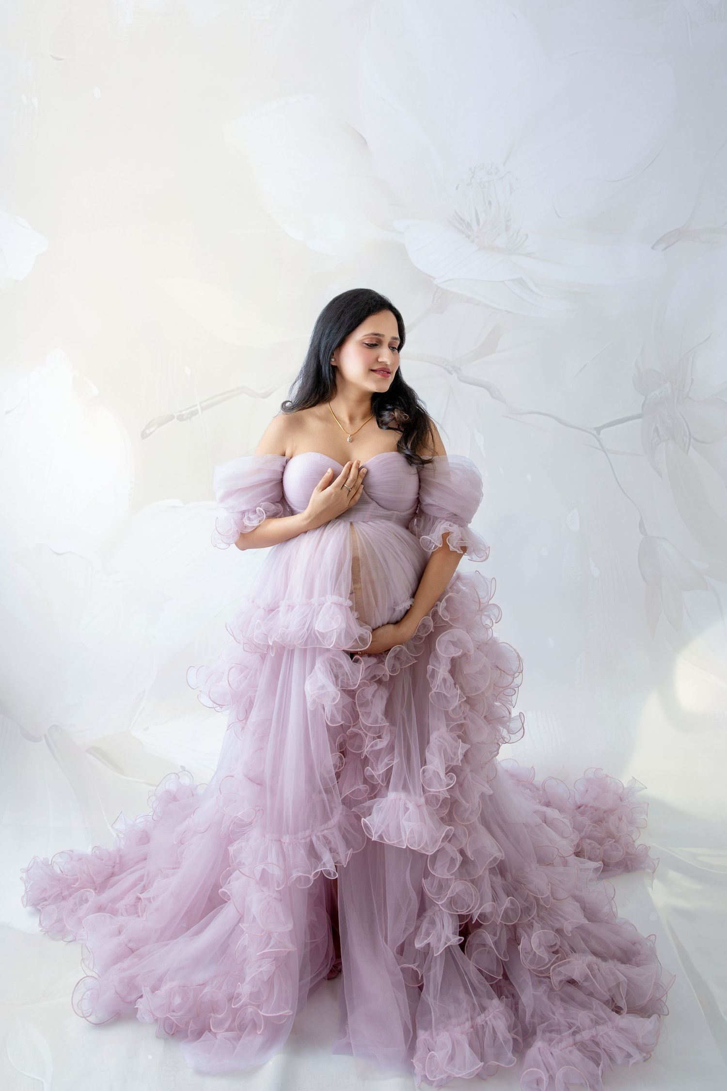 Pregnant woman in elegant lavender ruffle tulle gown posing gracefully against a soft floral white backdrop.