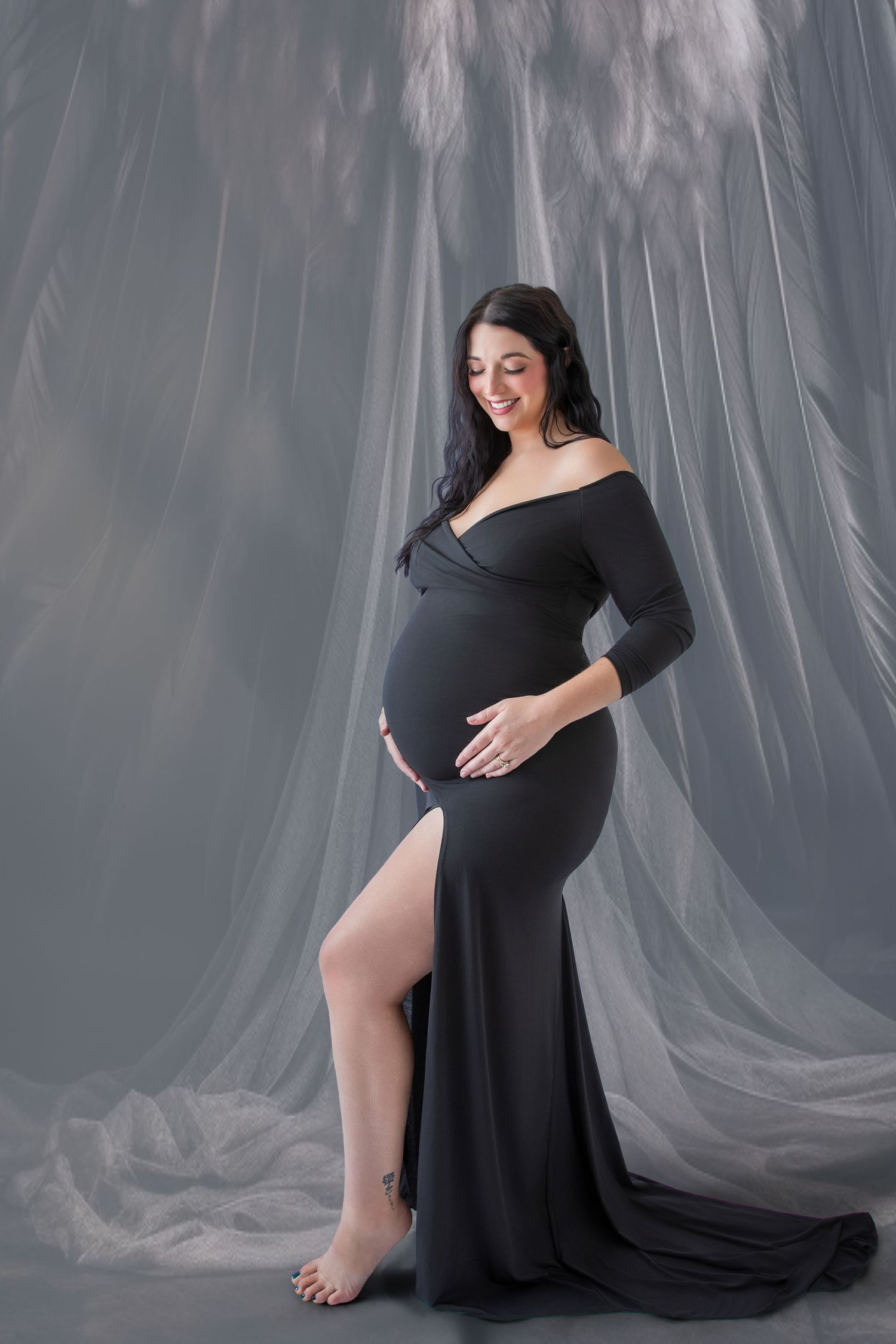Pregnant woman in elegant black off-shoulder gown with slit, posing in studio with flowing gray fabric backdrop.