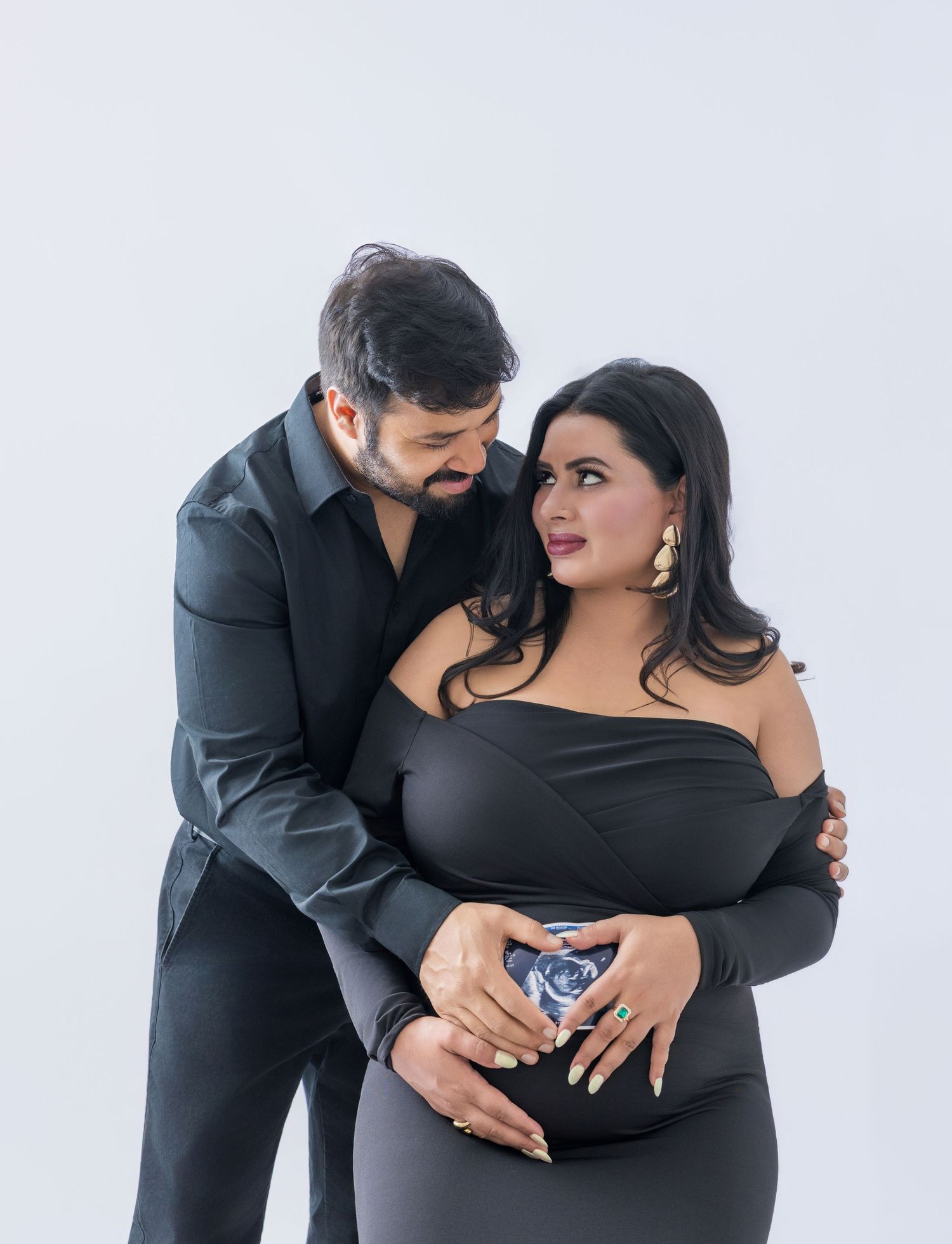 Couple in black outfits posing for maternity photo, woman holding ultrasound image over baby bump on white background.