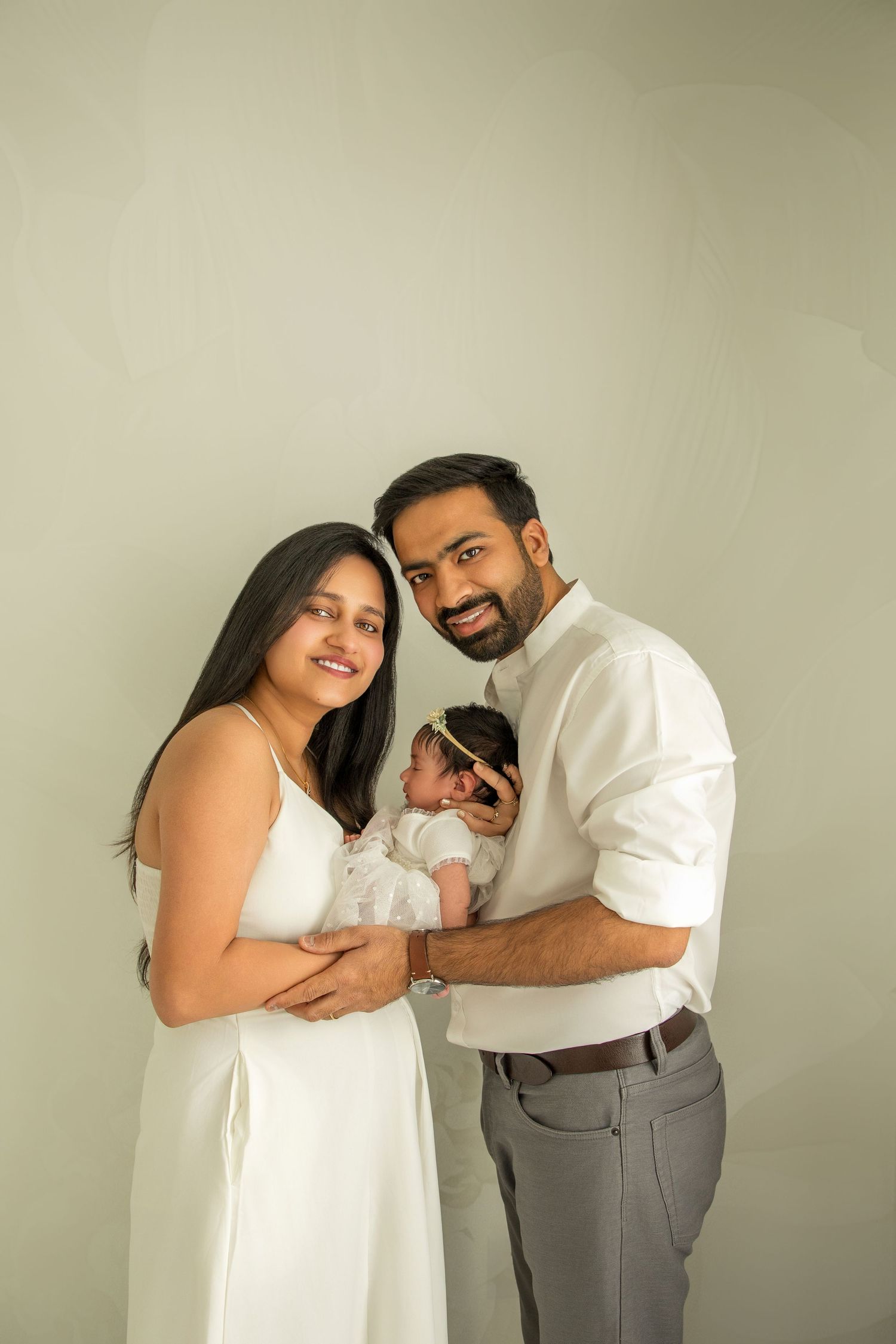 A couple in matching white and gray clothing embraces while holding their newborn during a professional photo session.