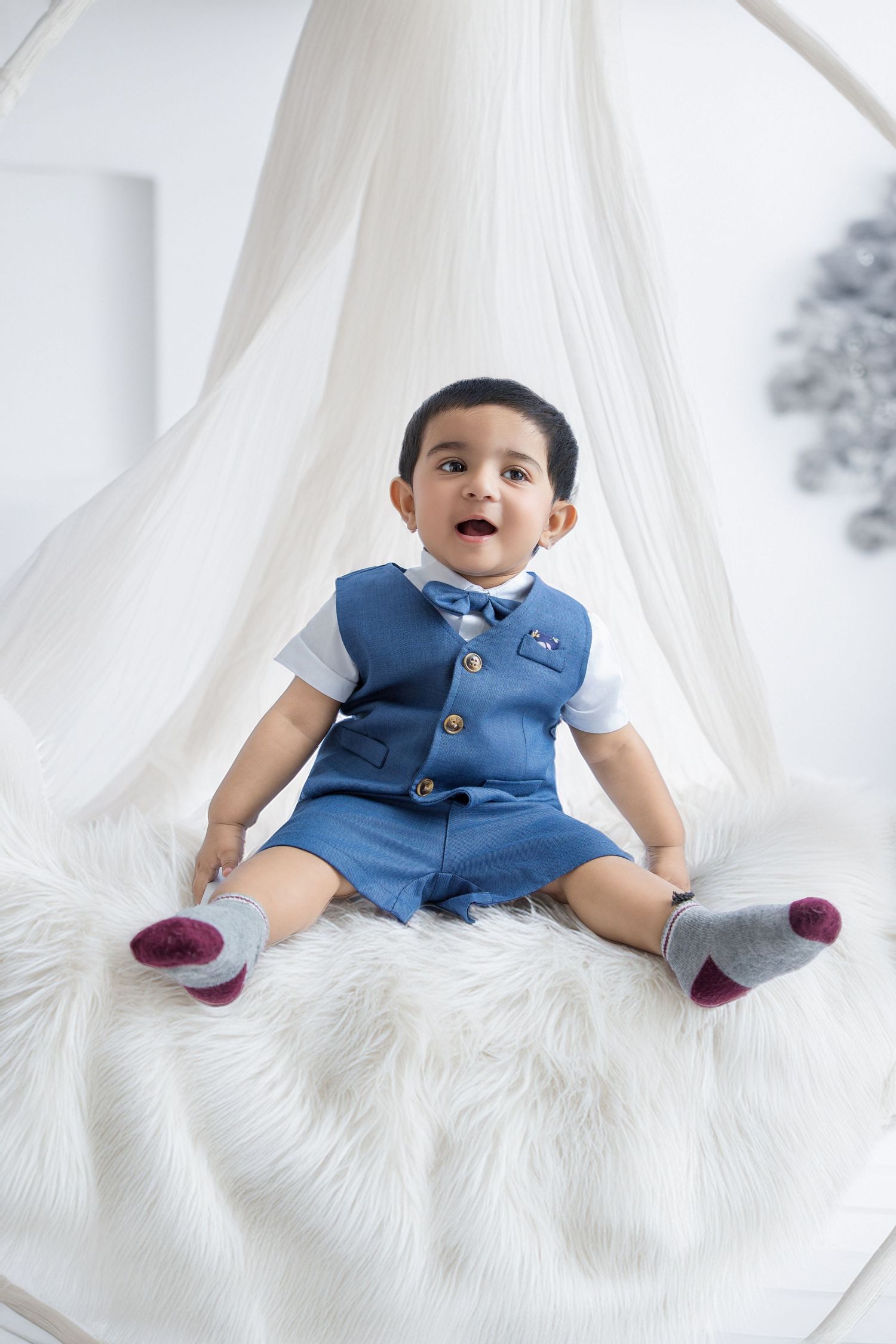 A baby in a blue vest and white shirt sits on a white fur rug in a bright studio setting.