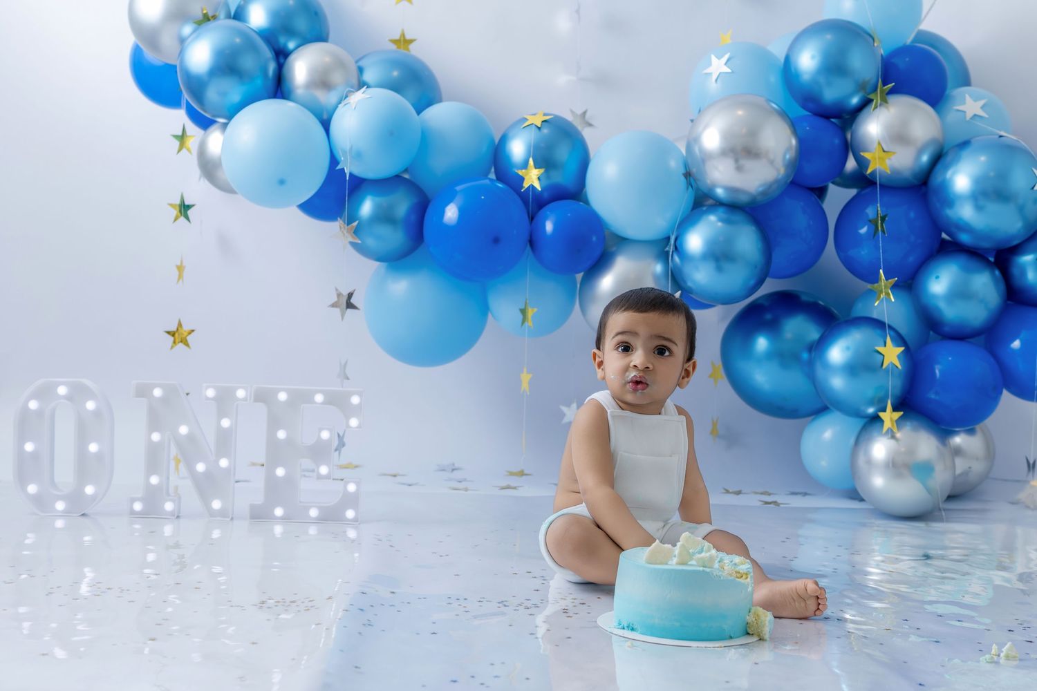 A festive first birthday celebration setup with blue and silver balloons and a cake against a light background.