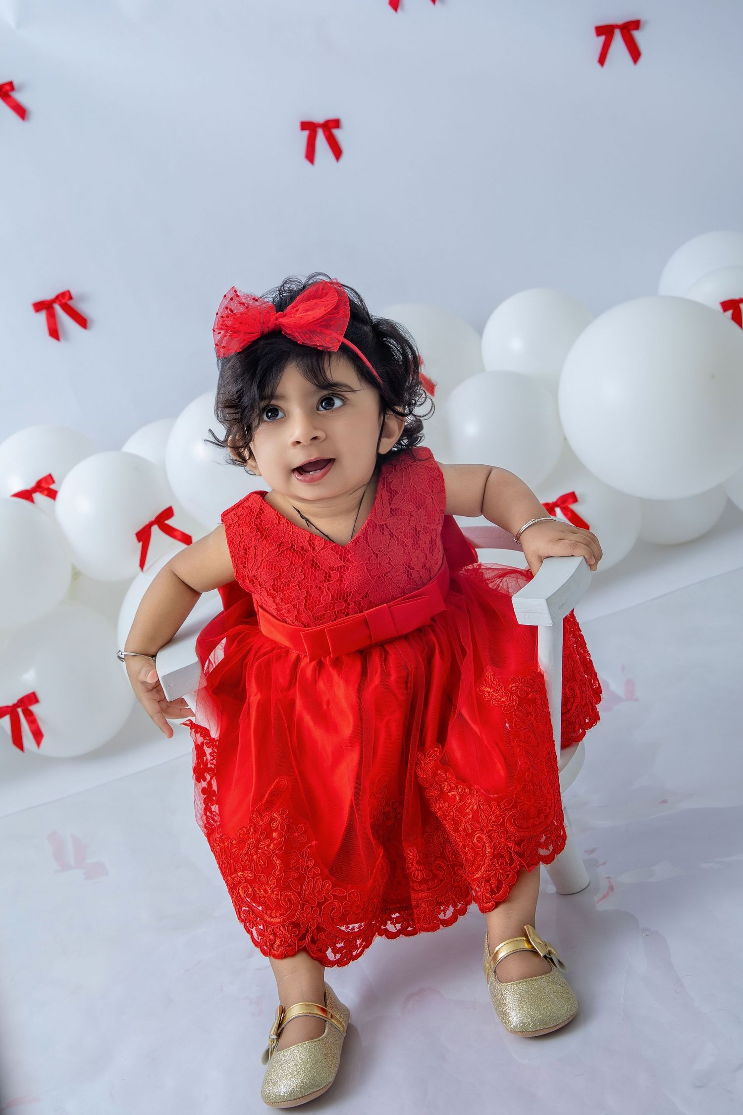 A child in a red lace dress and gold shoes explores the white studio setting.