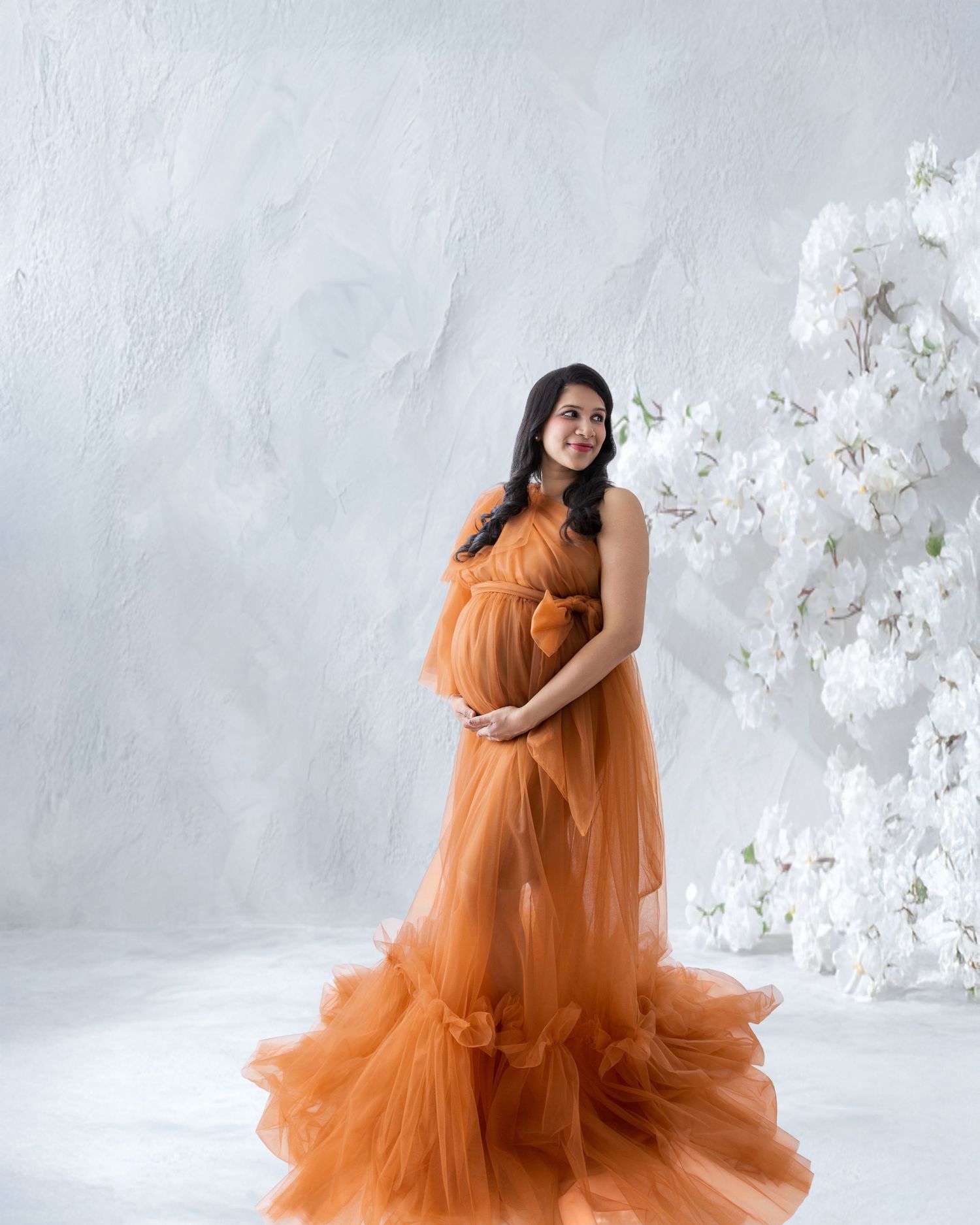 Pregnant woman in elegant orange tulle gown posing gracefully beside white floral arrangement in studio.
