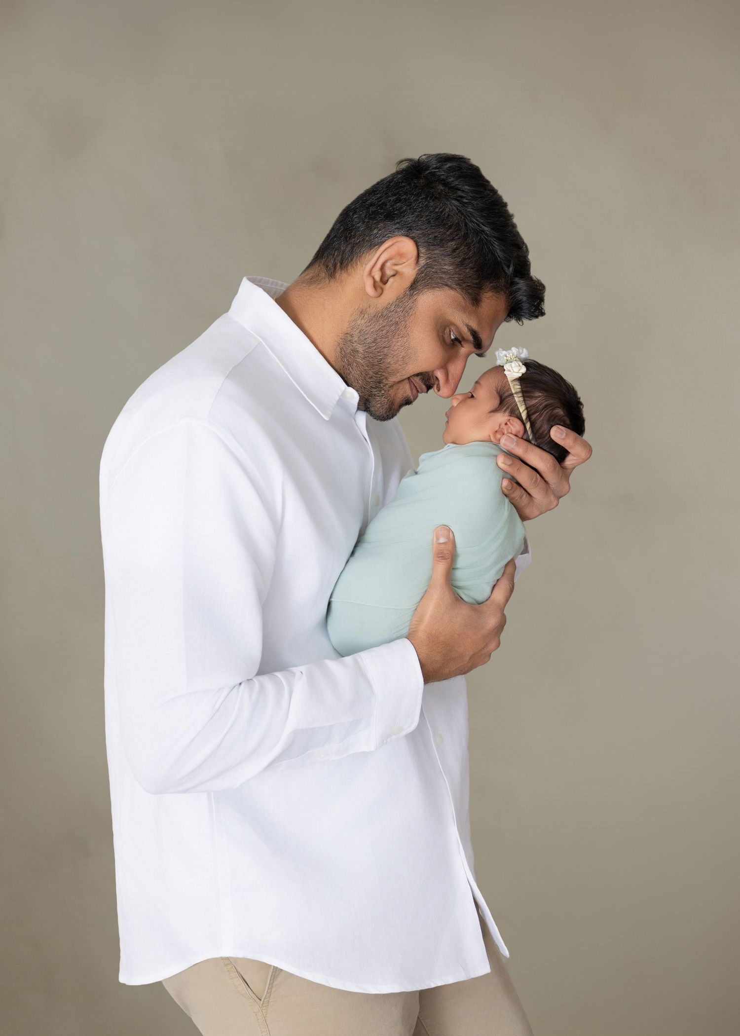 Father in white shirt tenderly holds swaddled newborn baby in mint green wrap against gray background.