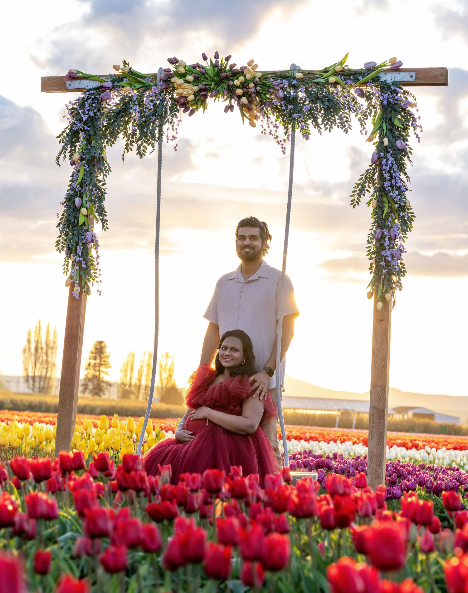 Couple poses under floral arch in tulip field at sunset, woman in red dress, man in white shirt.