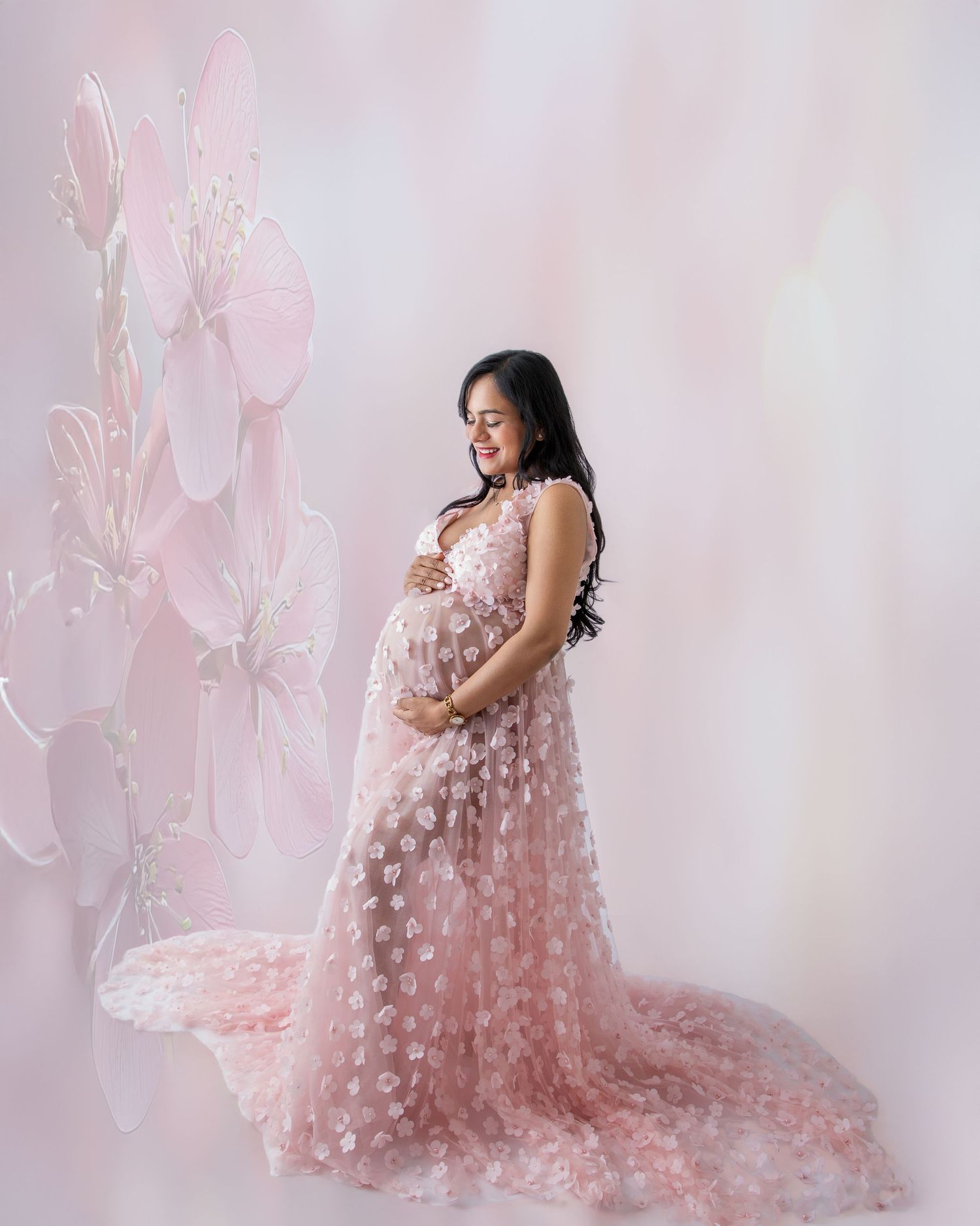 Pregnant woman in pink floral tulle maternity gown poses gracefully against a soft pink floral backdrop.