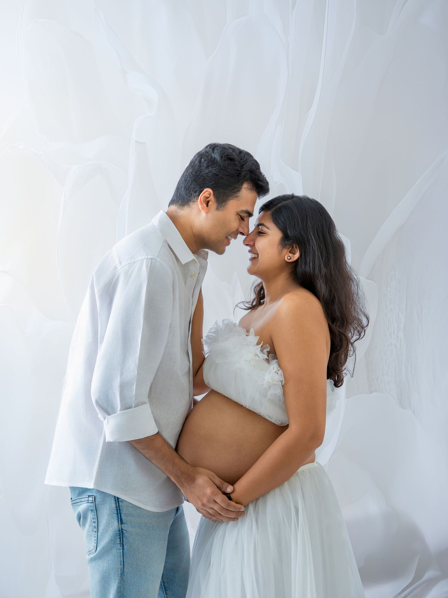 A couple in white outfits share a tender moment, the pregnant woman cradling her baby bump in a soft, ethereal studio setting.