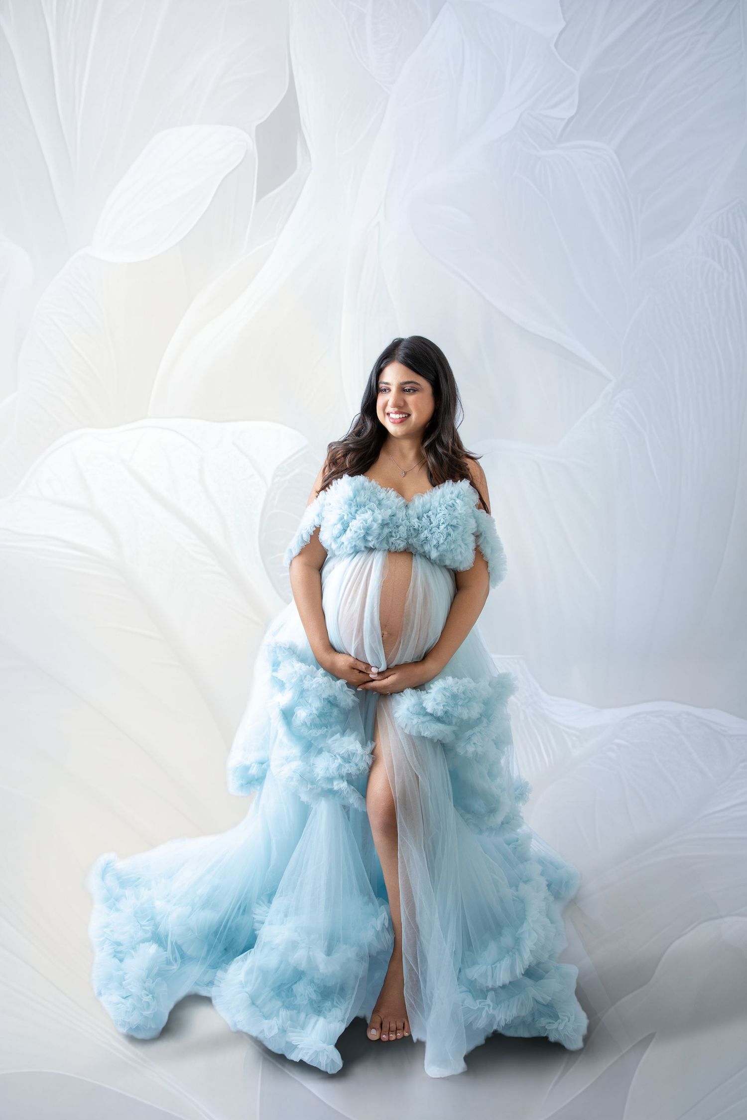 Pregnant woman in a flowing light blue ruffle tulle gown poses gracefully in a bright studio maternity photoshoot.