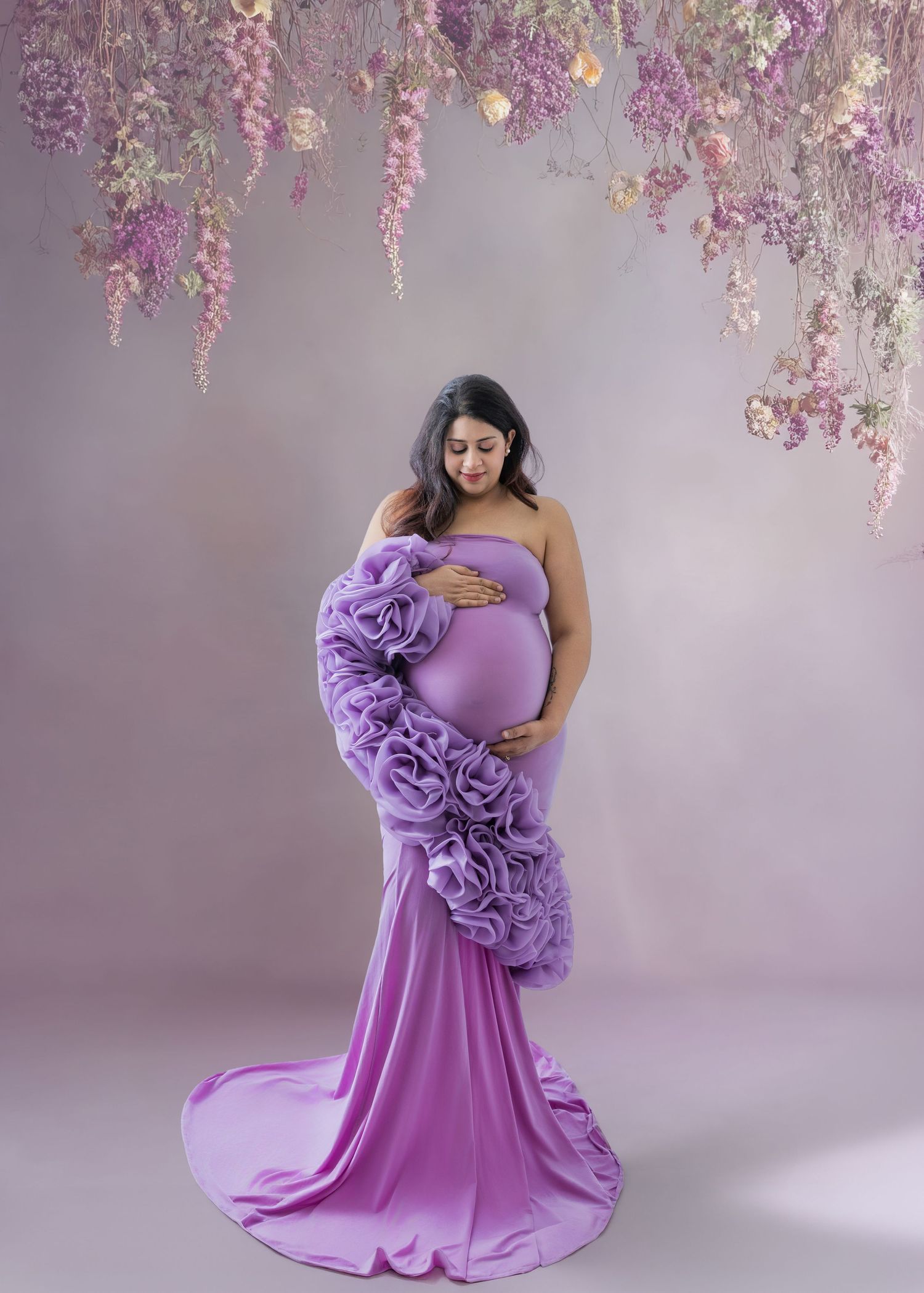 Pregnant woman in purple ruffle maternity gown poses under hanging flowers in a dreamy studio portrait.