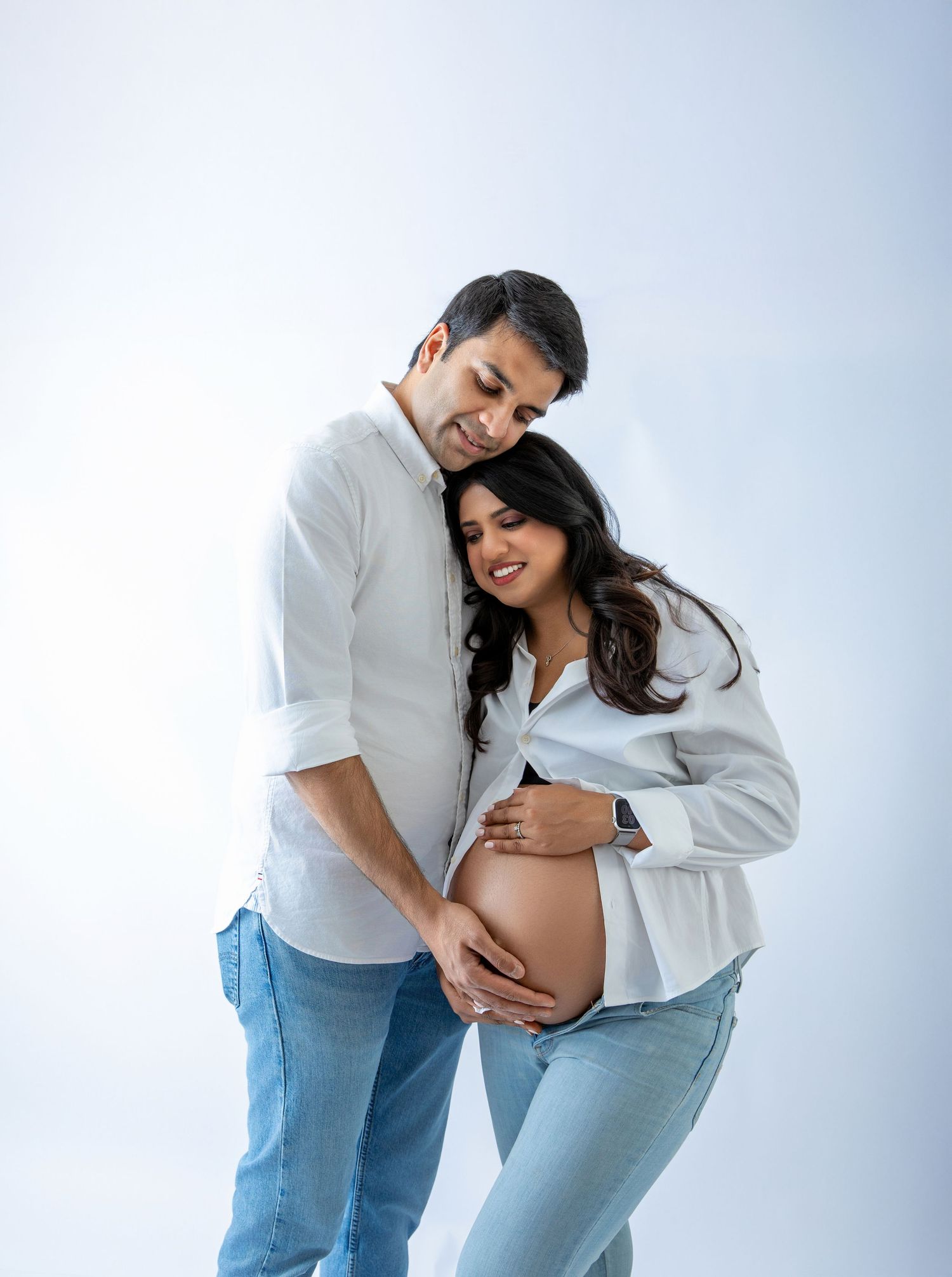 Couple in matching white shirts and jeans posing for a maternity photo shoot on a white background.