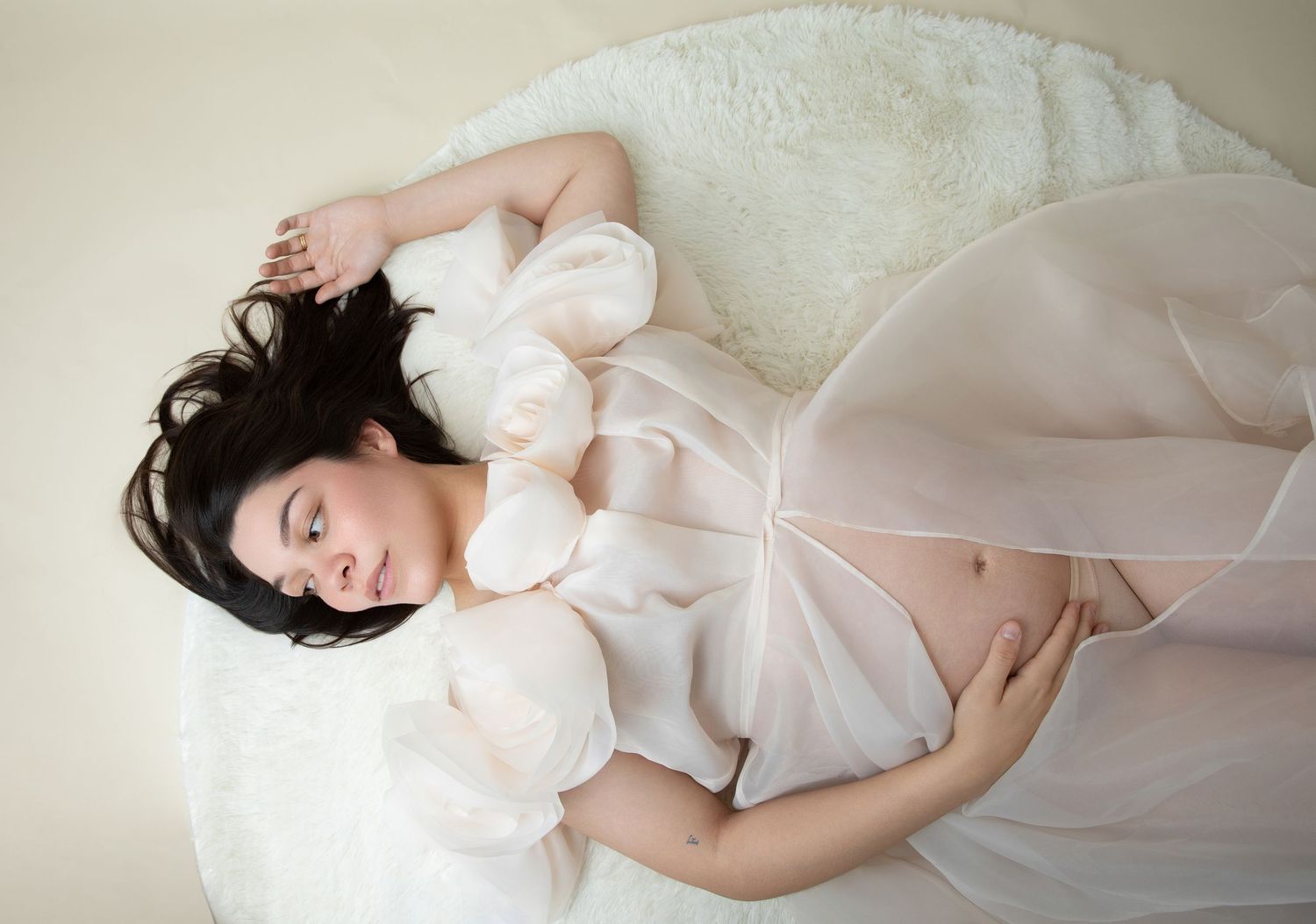 Pregnant woman in white chiffon gown lying gracefully, cradling baby bump in a soft, dreamy maternity photo shoot.