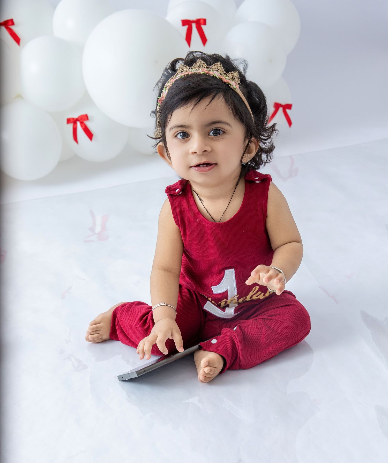 A toddler in a red outfit sits on white floor with decorative red bows in the background.