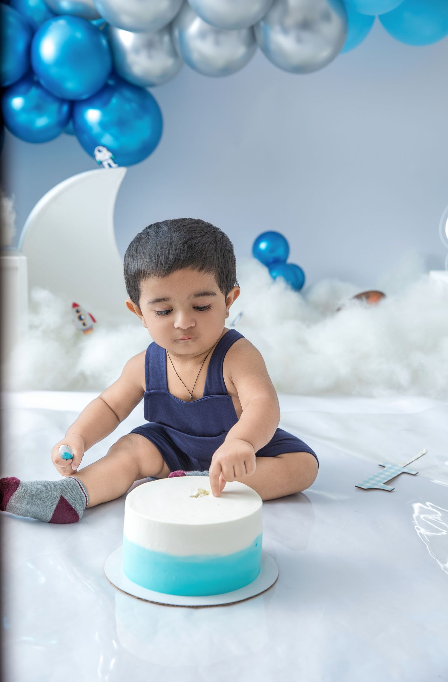 Child in navy sleeveless outfit explores birthday cake with blue and silver balloon decorations.