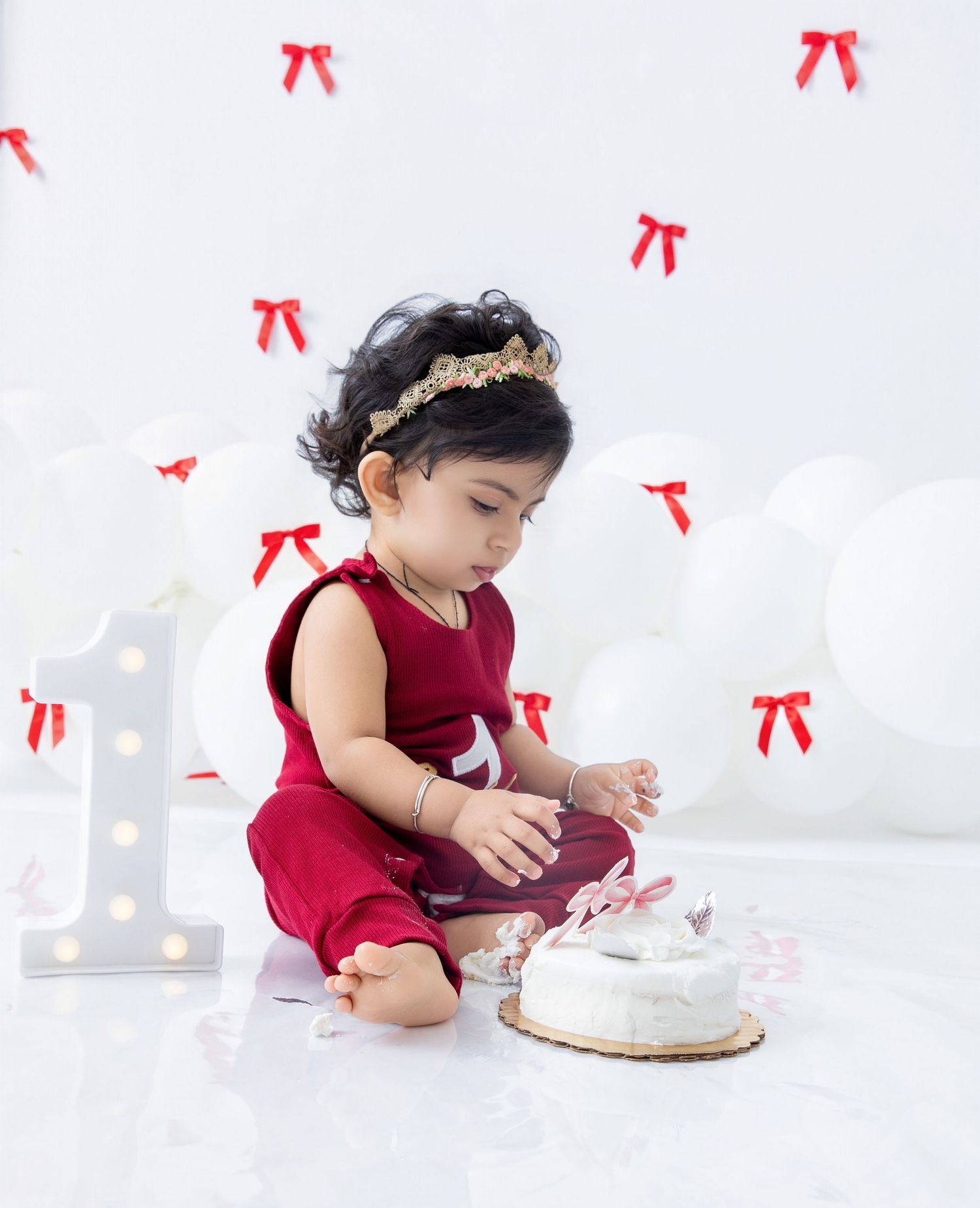 A small child in red clothing reaches toward a white cake against a light background with red bows.