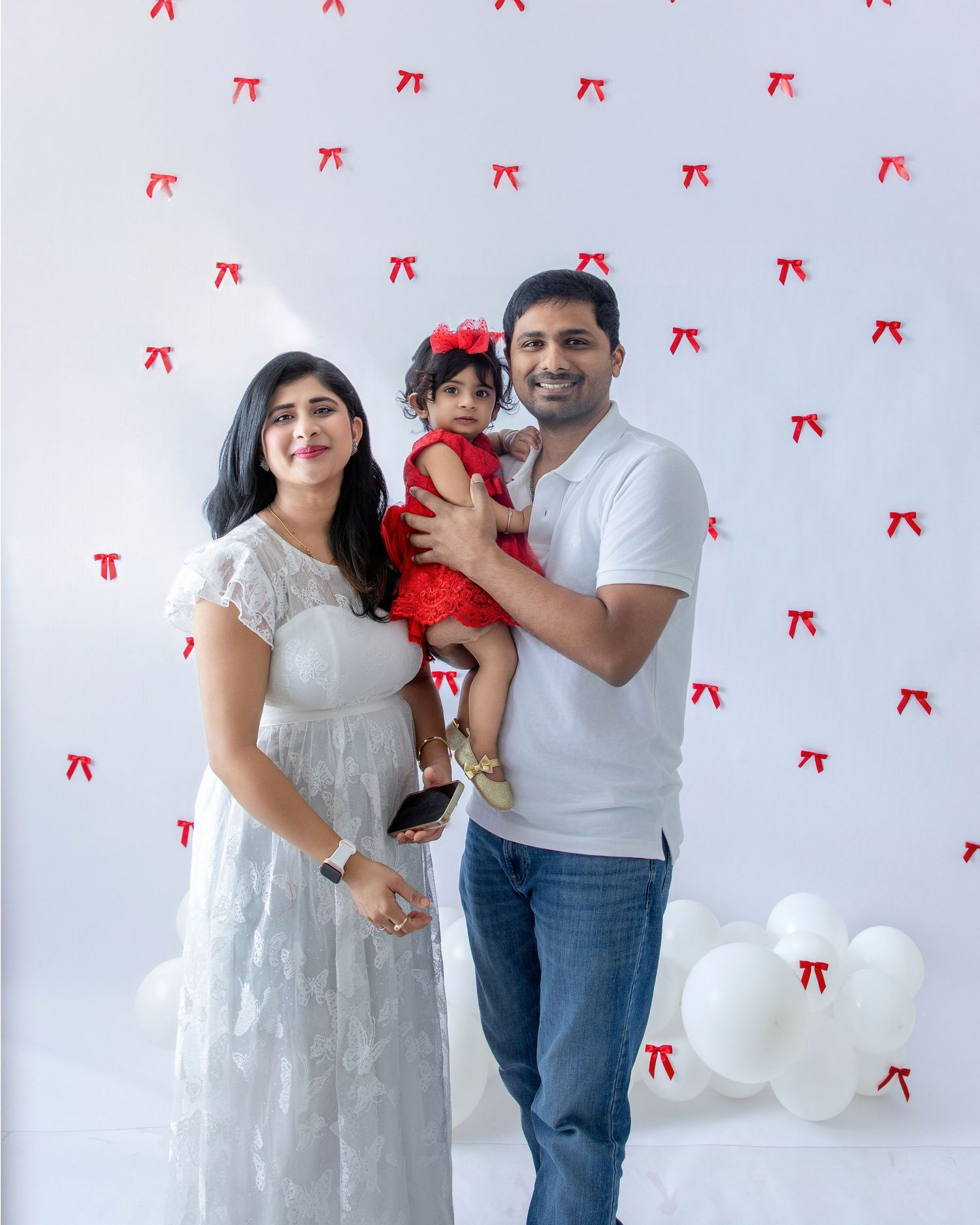 Family in white and red outfits poses together against wall decorated with red bows.