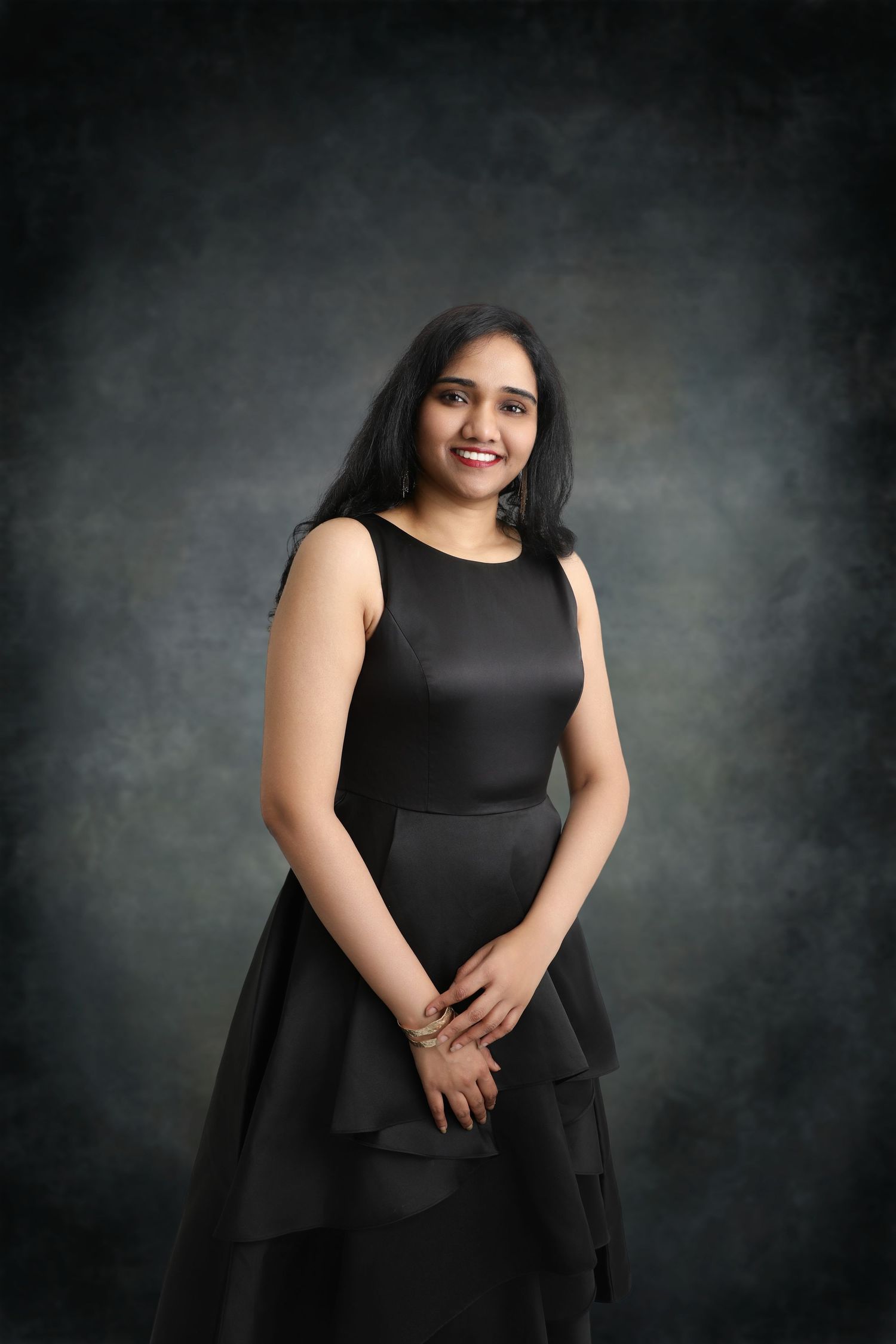 Woman in elegant black dress poses against dark dramatic background.