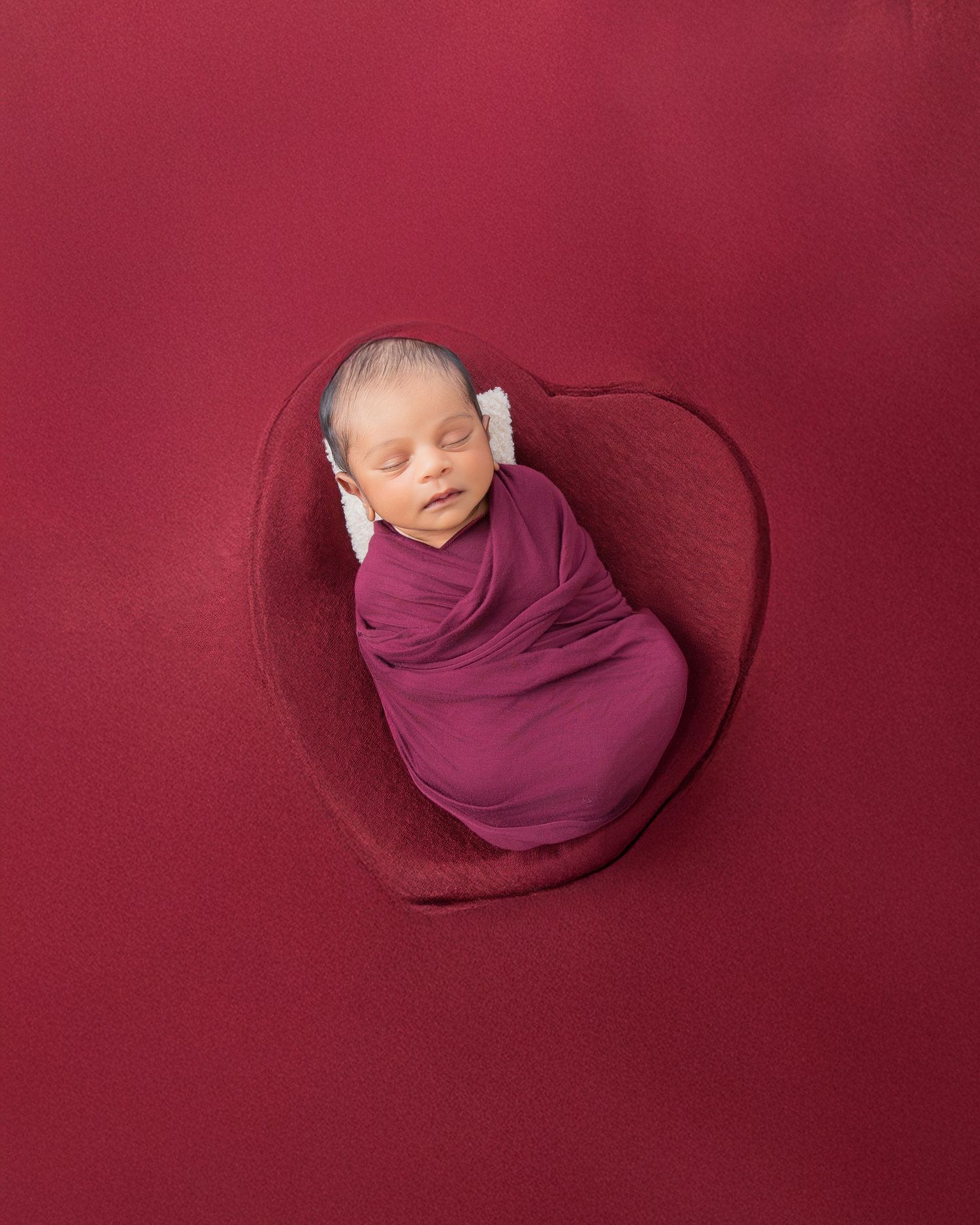 A newborn baby wrapped in a burgundy swaddle blanket lies peacefully on a matching red textured backdrop.