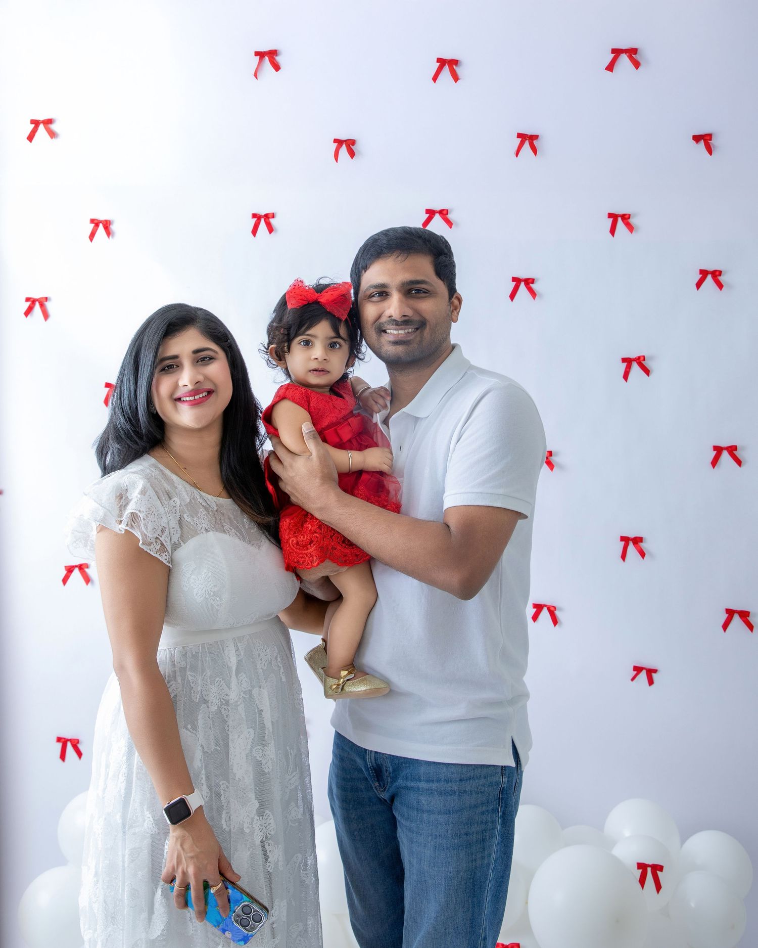 Family poses together against white backdrop with red bow decorations and white balloons.